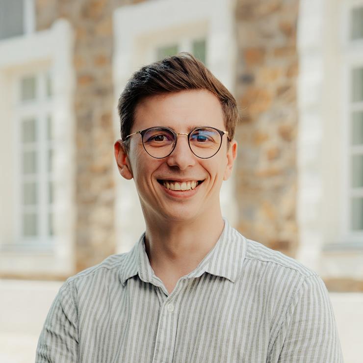 Junger Mensch mit Brille und gestreiftem Hemd lächelt draußen vor einem Gebäude.