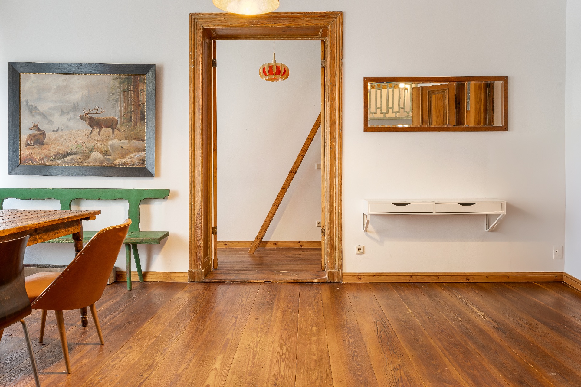 Wohnzimmer mit Holzboden, Tisch, grüner Bank, Wandbild, Spiegel und offenem Türrahmen.