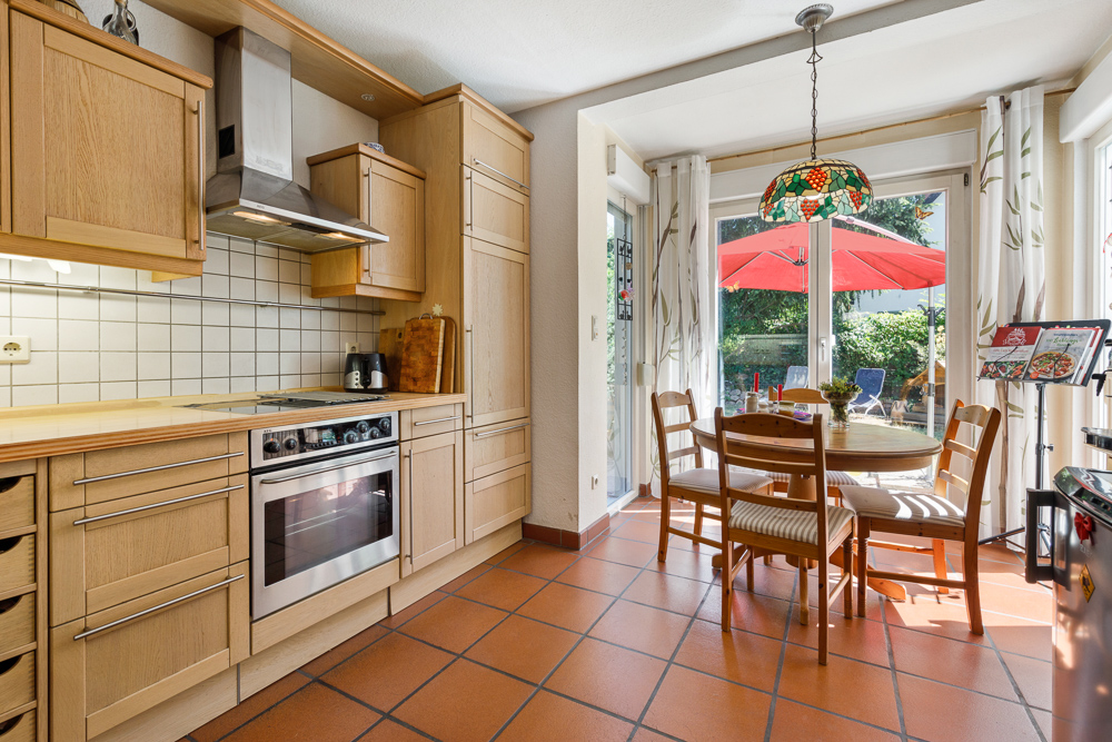 Helle Küche mit Holzmöbeln, Esstisch am Fenster und Zugang zur Terrasse mit rotem Sonnenschirm.