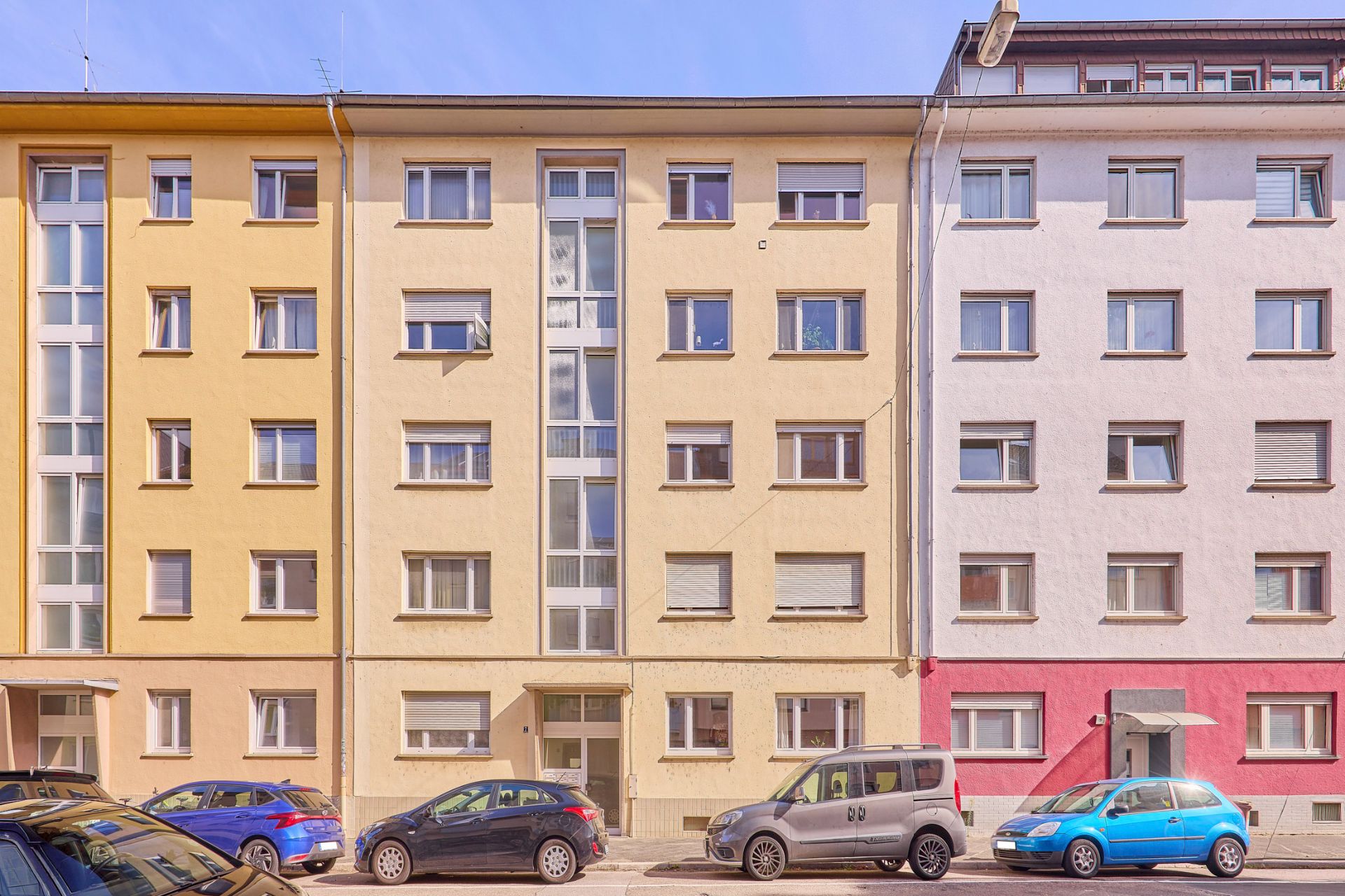 Mehrstöckiges Wohnhaus mit gelber und beiger Fassade, mehrere Autos parken davor.