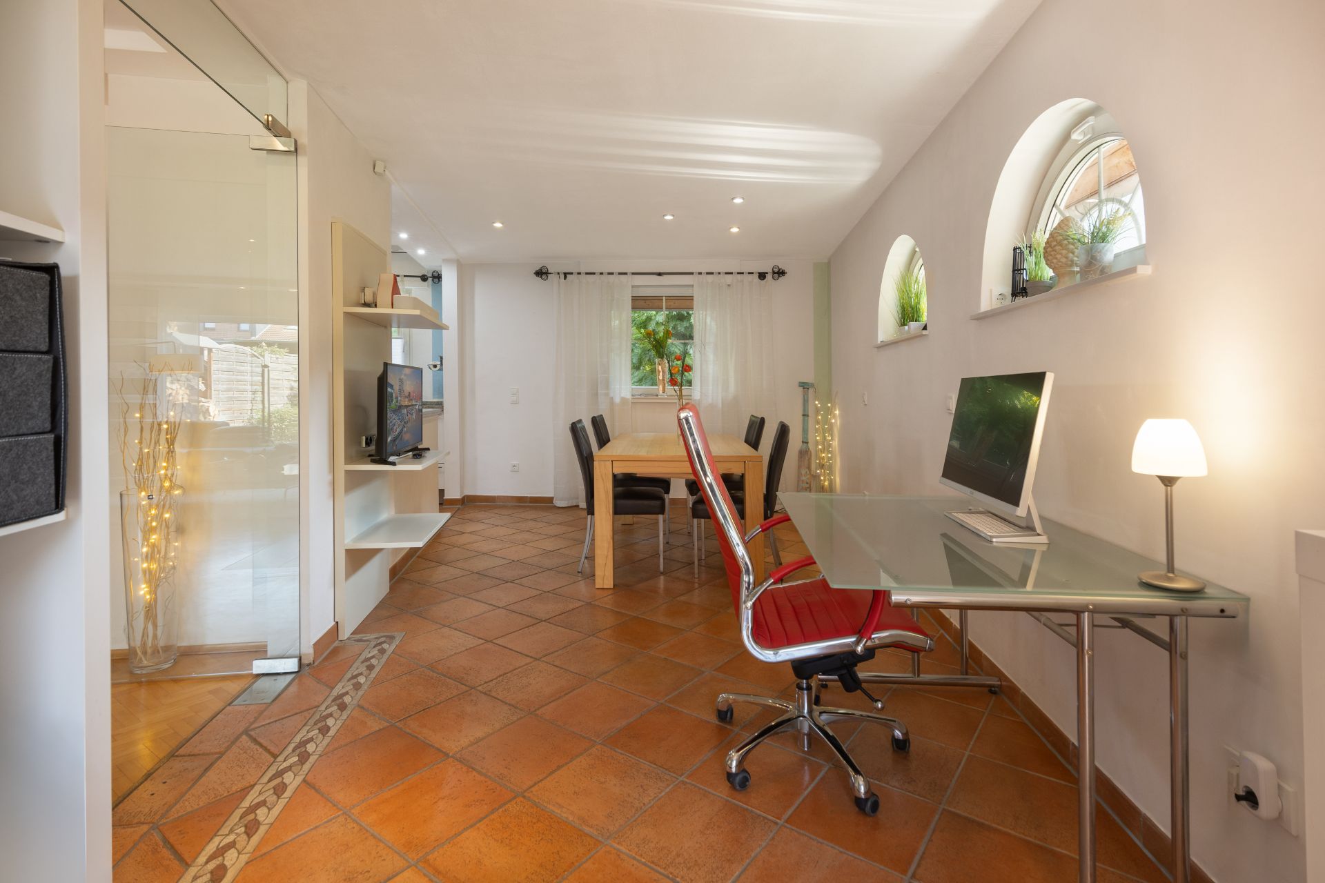 Helles Arbeits- und Esszimmer mit Glastisch, Stuhl, Esstisch und Fenstern mit Pflanzen.