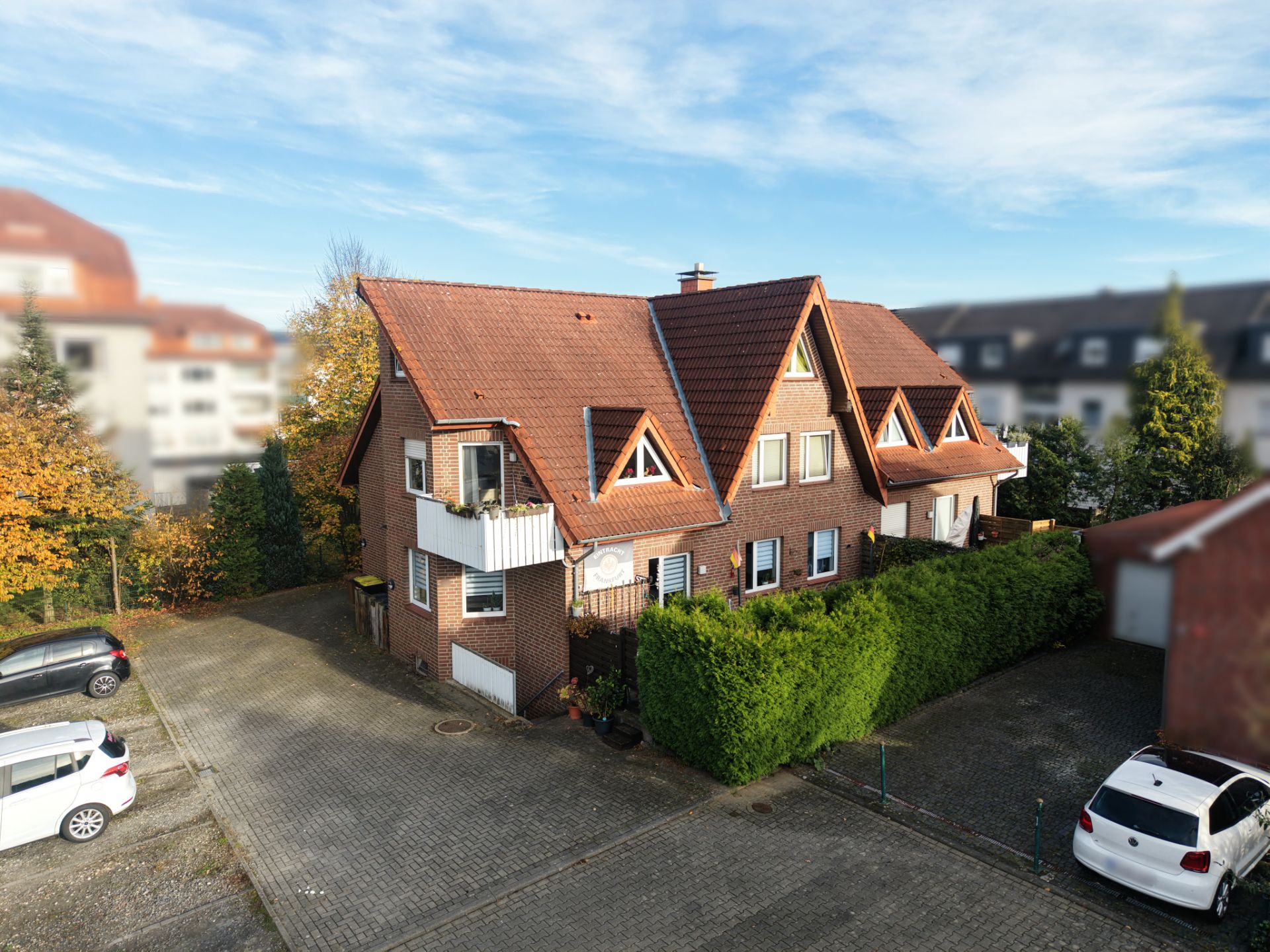 Backsteinhaus mit rotem Dach, Balkon und umliegendem Parkplatz, daneben grüne Hecke.