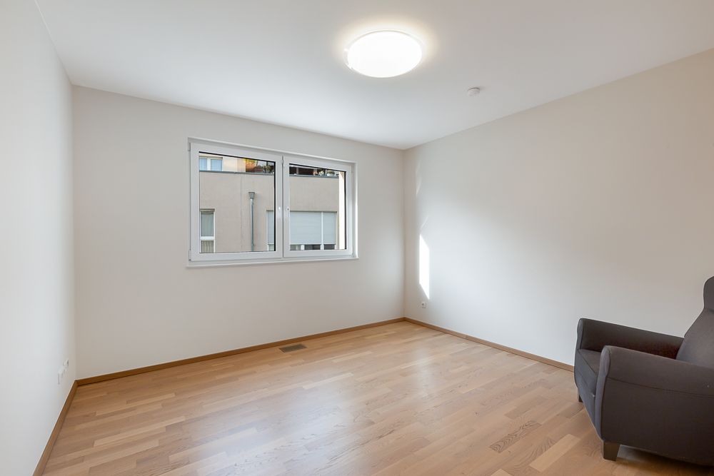 Helles Zimmer mit Holzfußboden, großem Fenster und einem einzelnen Sessel in der Ecke.