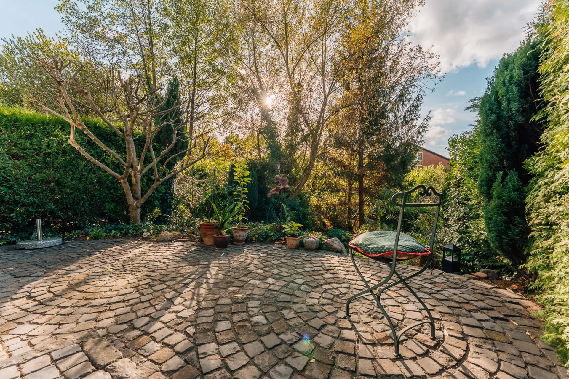 Gepflasterte Terrasse mit Stuhl, Blumentöpfen und Bäumen im Garten bei Sonnenschein.