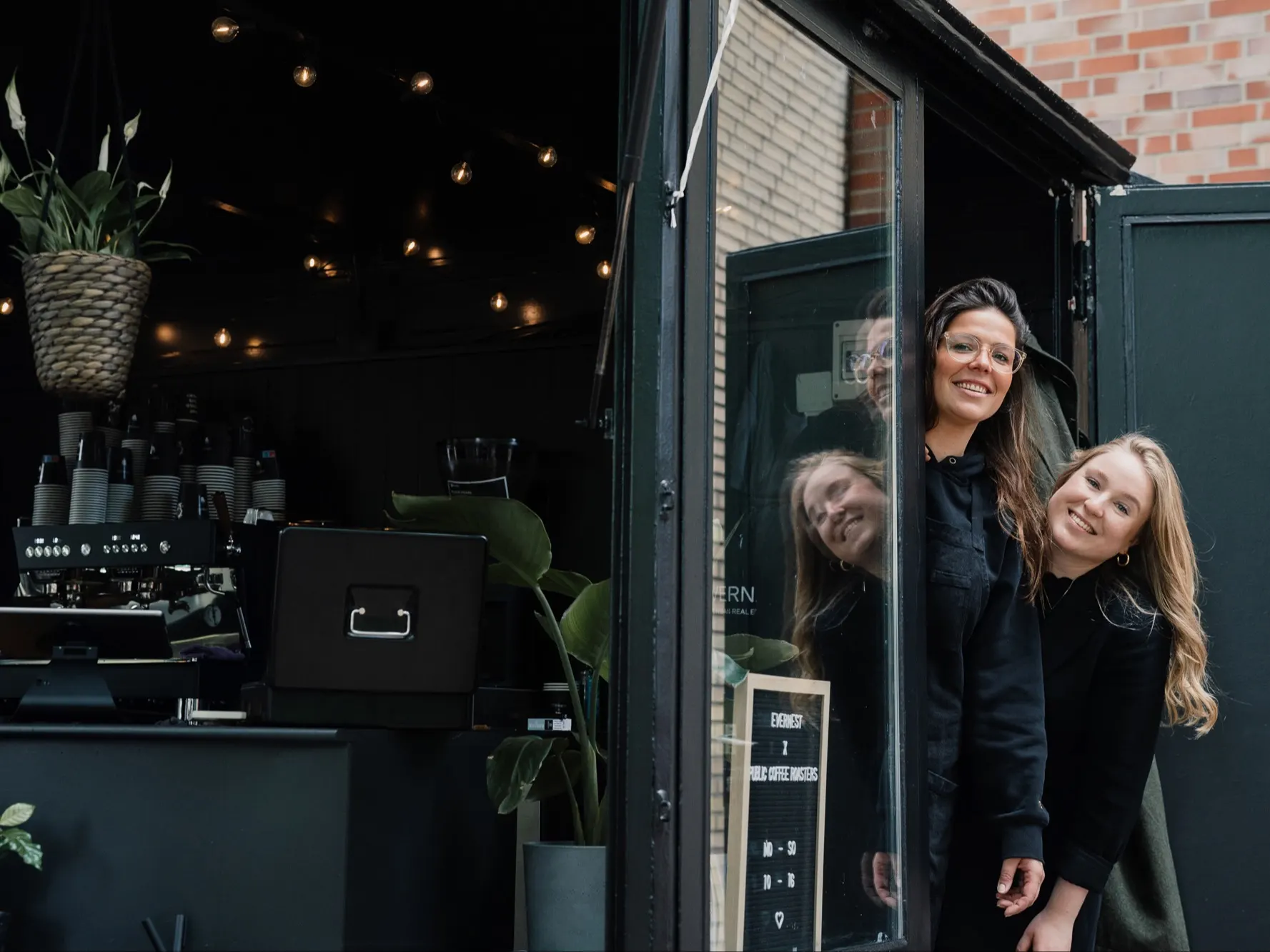 Zwei Frauen stehen lächelnd an der offenen Tür eines kleinen Cafés mit Pflanzen und Lichterkette.