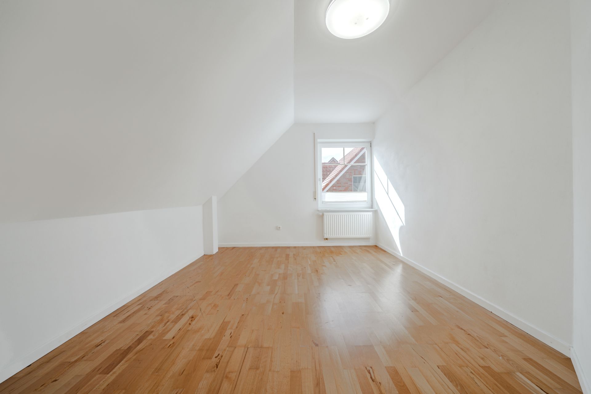 Heller Raum mit schräger Decke, Holzfußboden und einem Fenster mit Heizkörper darunter.
