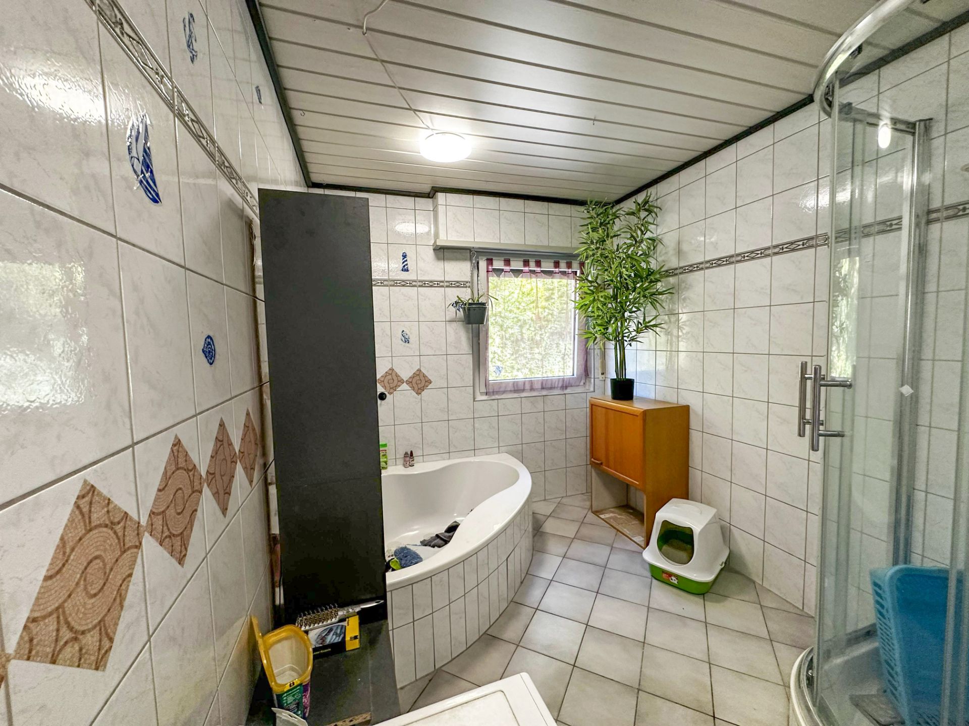 Gefliestes Badezimmer mit Eckbadewanne, Dusche, Fenster und Katzentoilette.