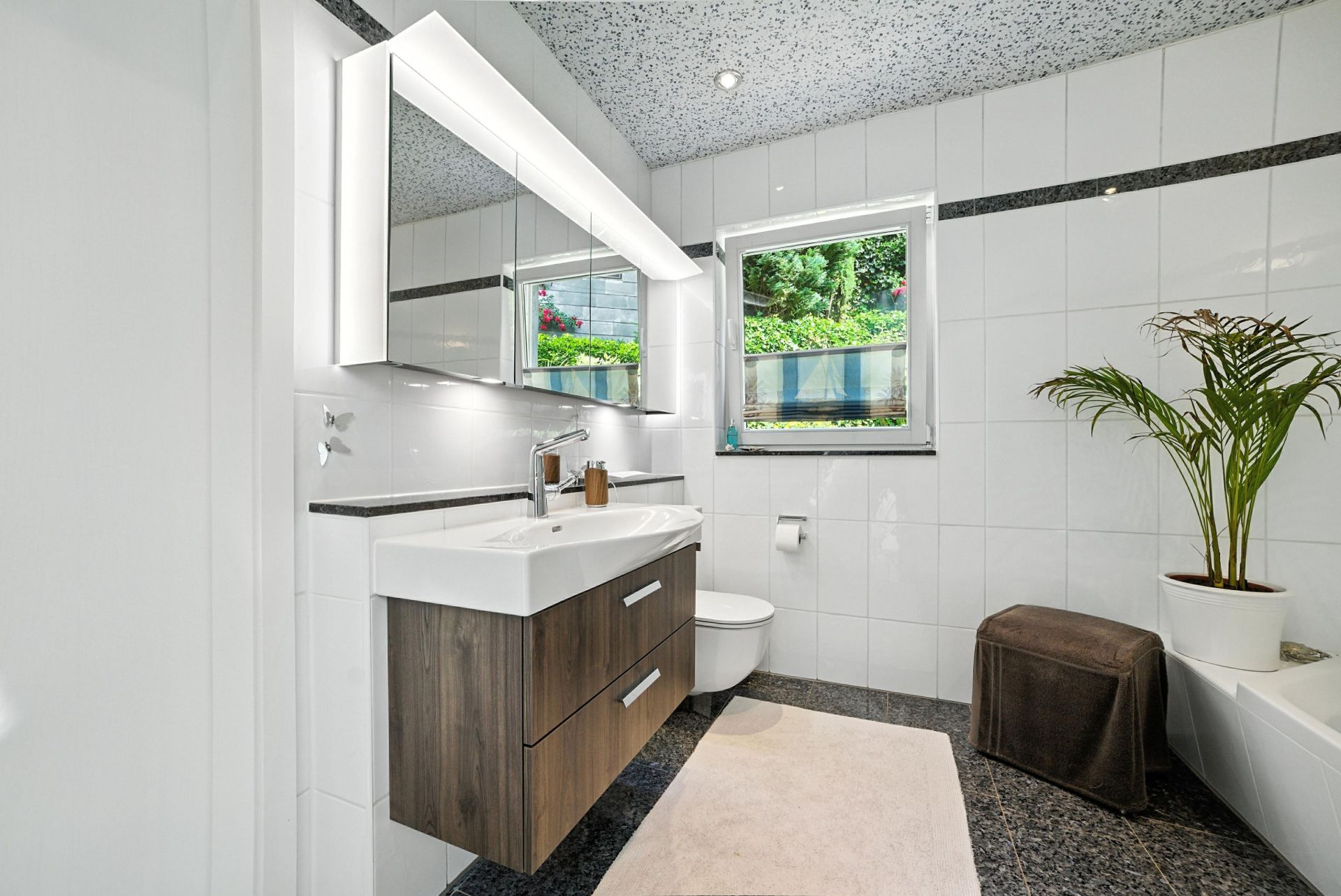Modernes Badezimmer mit großem Spiegel, Waschtisch, WC, Fenster, Pflanze und Hocker.
