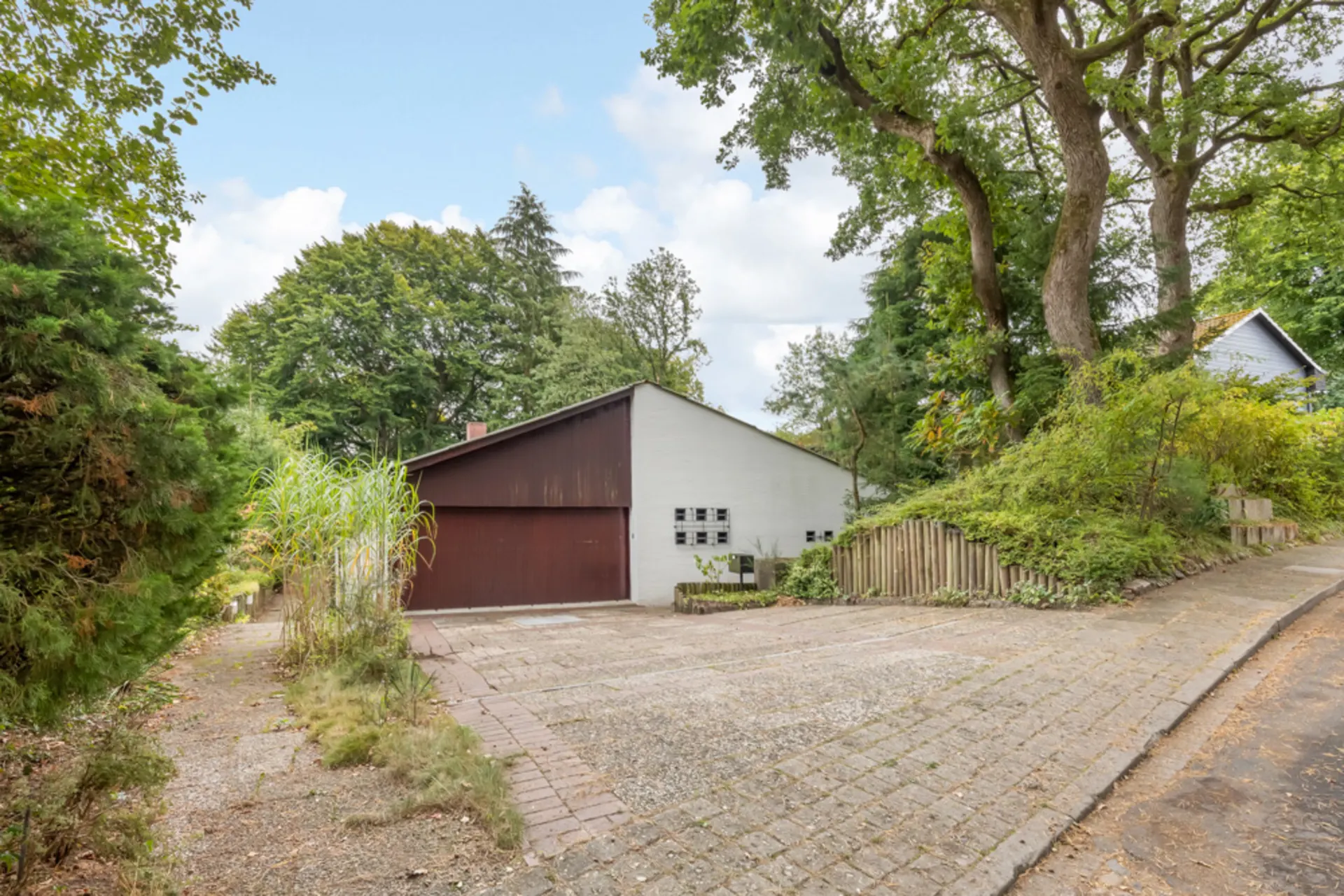 Garage mit braunem Tor und umgebenden Bäumen, gepflasterte Zufahrt im Vordergrund.