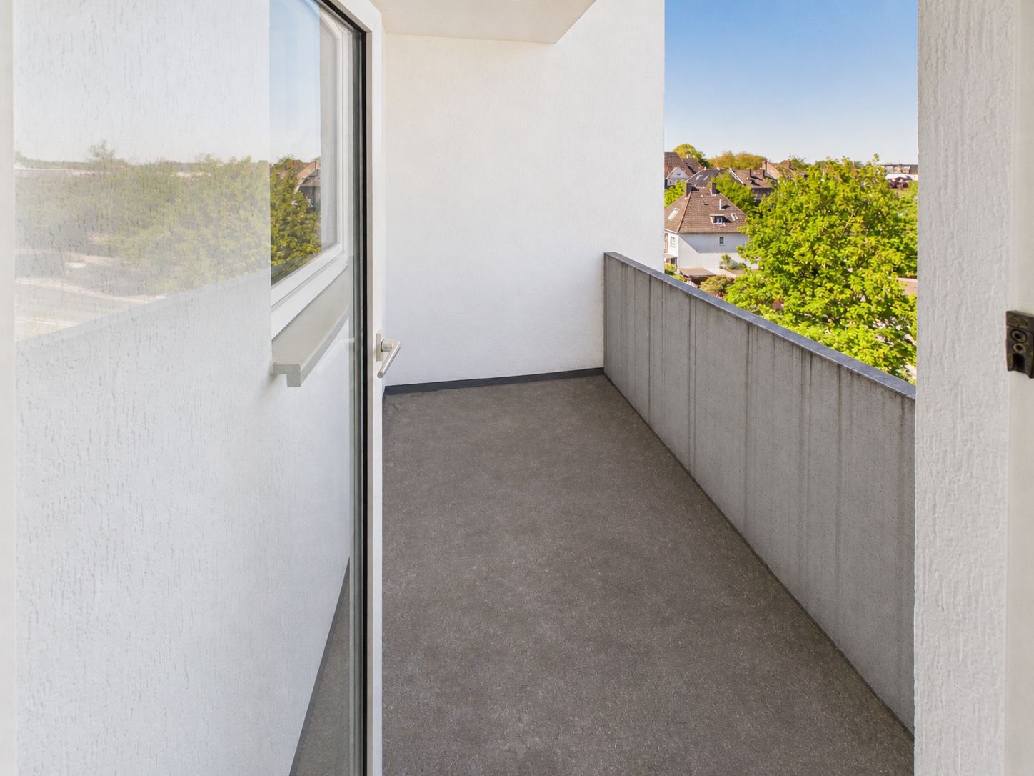 Moderner Balkon mit Betonboden und Blick ins Grüne, Zugang durch Glastür.