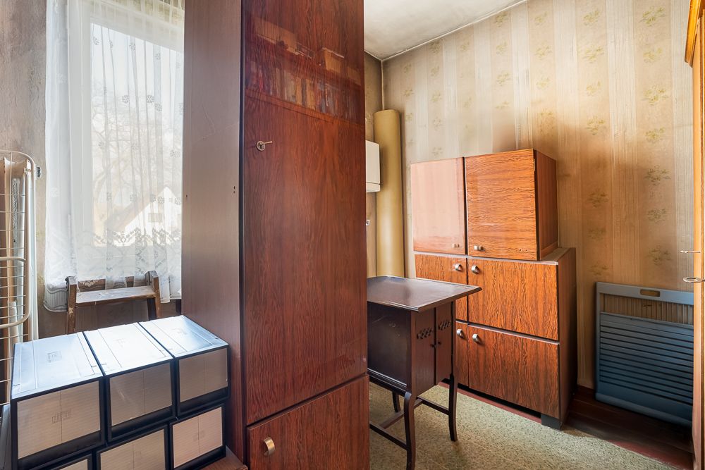 Kleines Zimmer mit alten Holzschränken, Schreibtisch und Kartons vor einem Fenster mit Gardine.