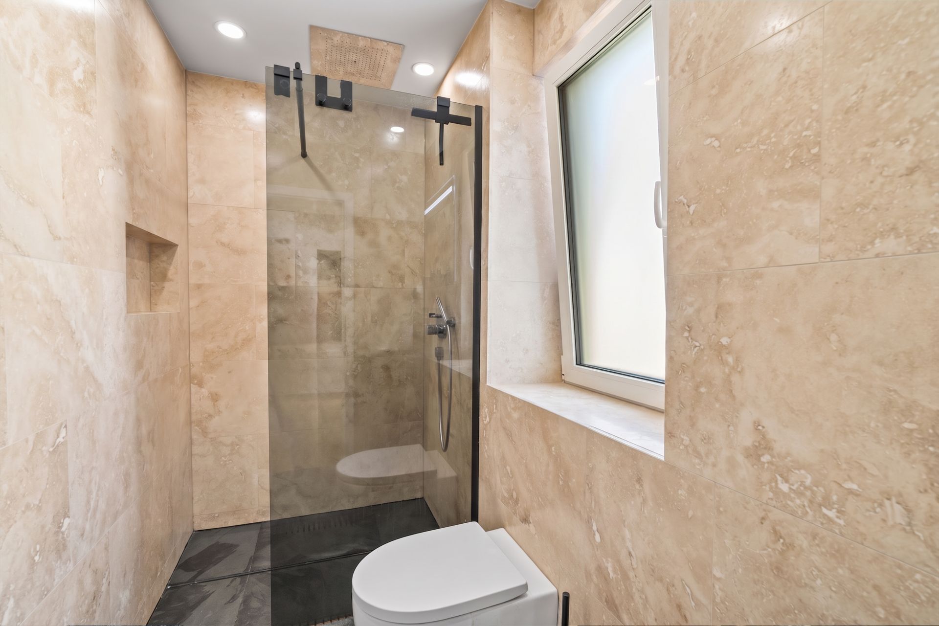 Modernes Bad mit großer Dusche, Glasabtrennung, Toilette und Fenster, beige gefliest.