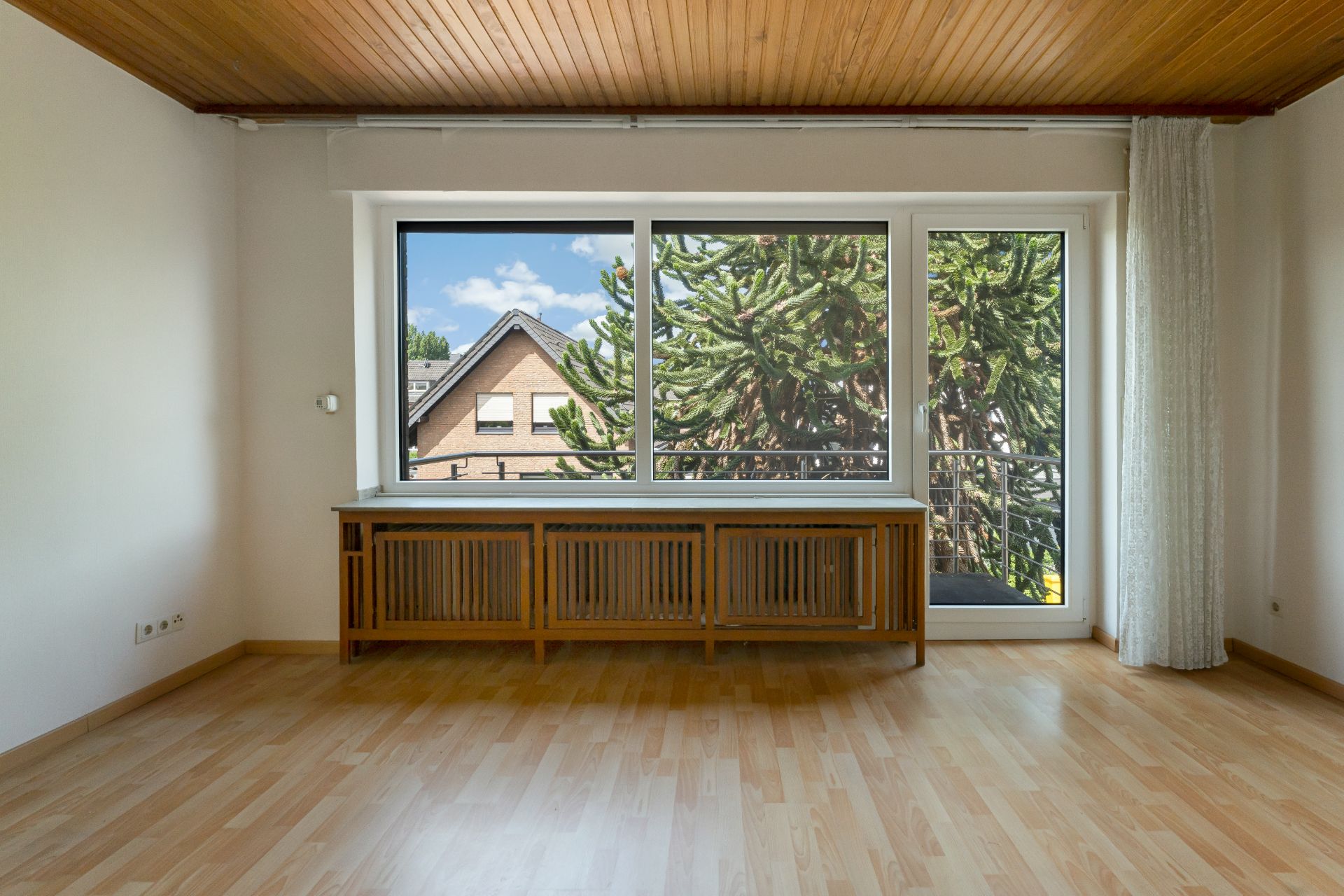Leeres Zimmer mit Holzdecke, Parkettboden, großem Fenster und Heizkörperabdeckung aus Holz.