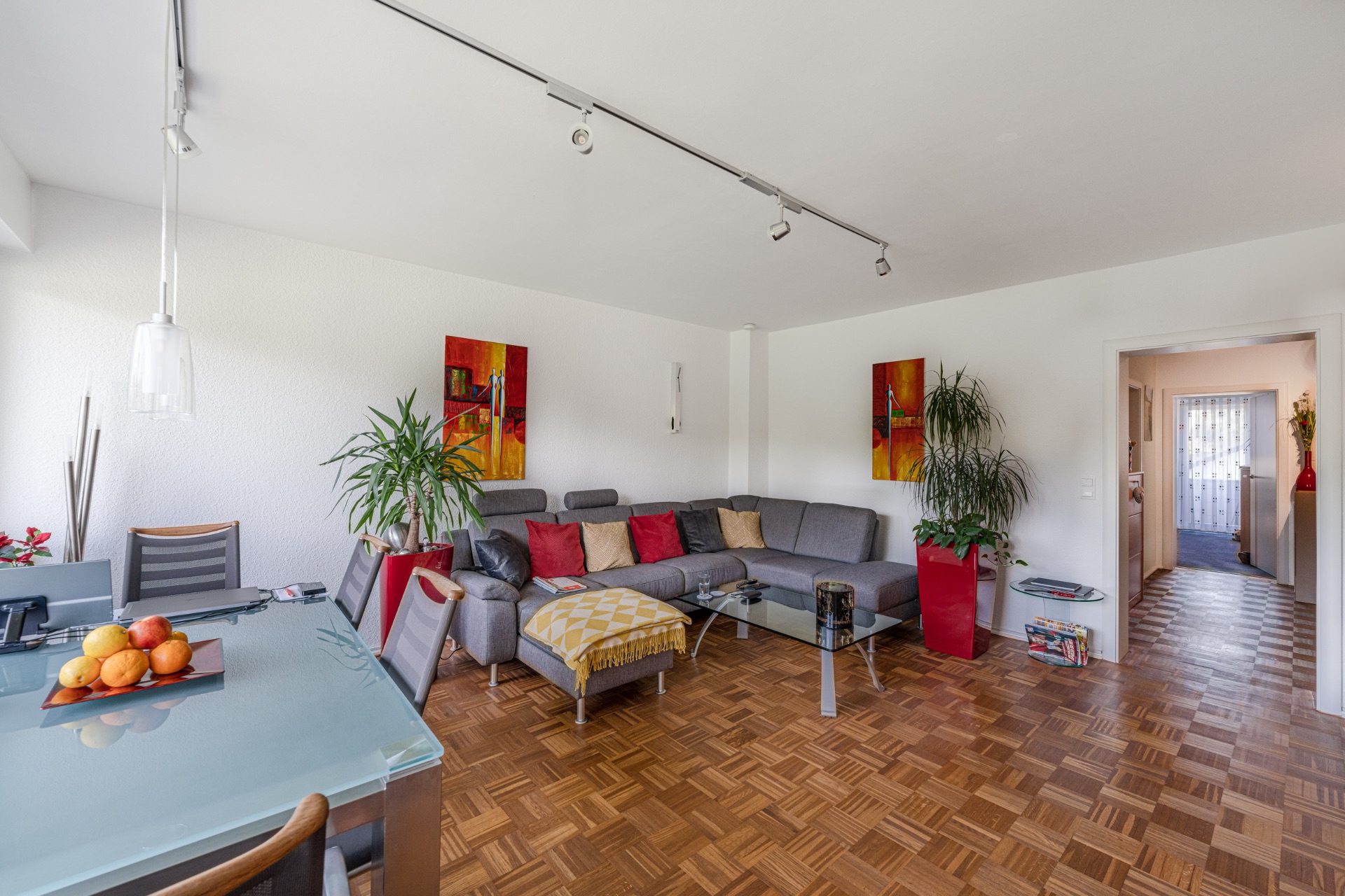 Modernes Wohnzimmer mit grauem Ecksofa, Glastisch, Esstisch und Pflanzen auf Parkettboden.