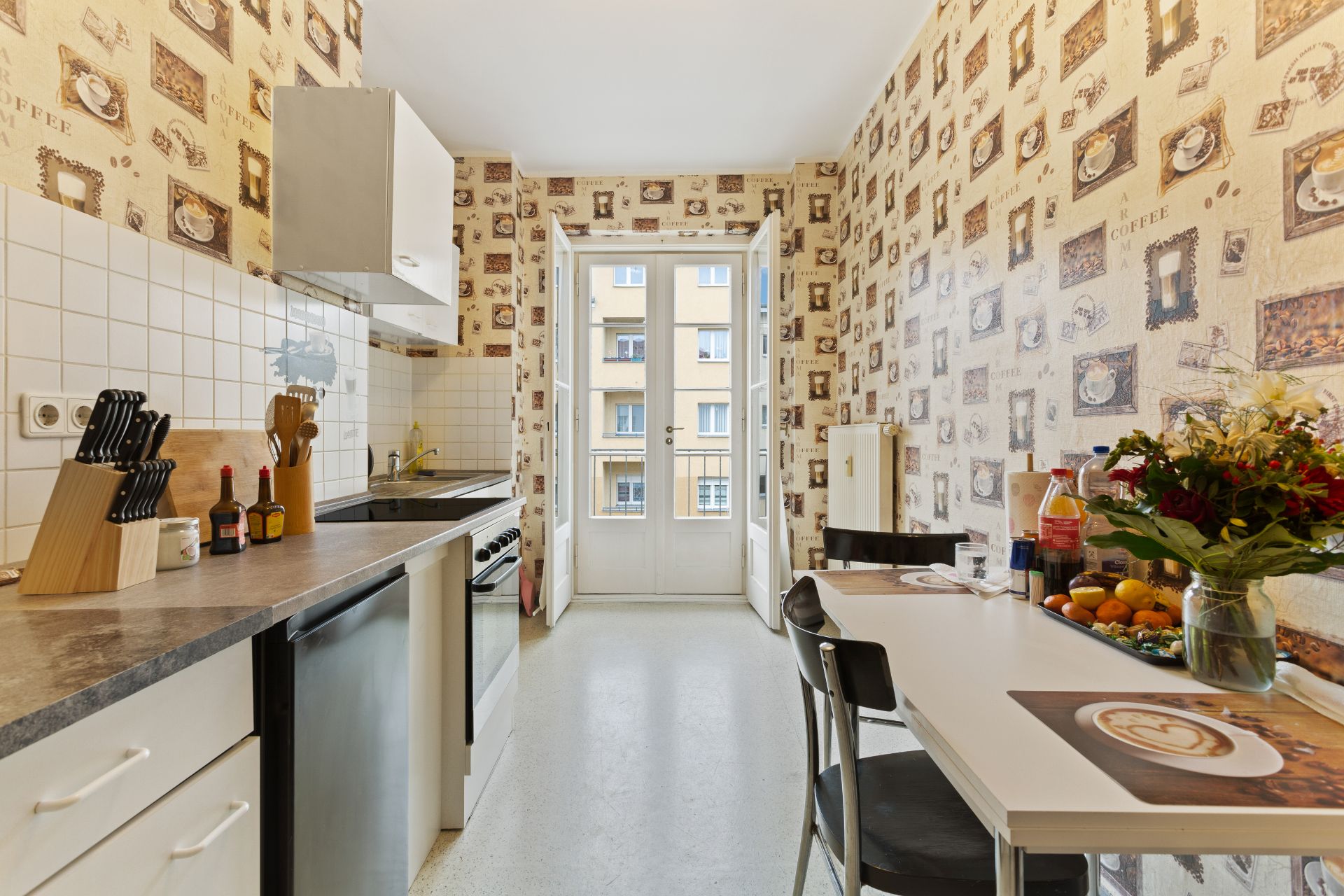 Helle Küche mit gemusterter Tapete, Esstisch, weißen Schränken und Glastür zum Balkon.