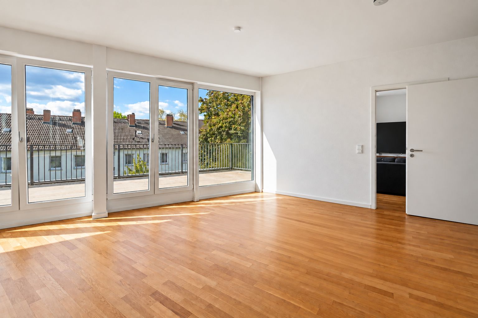 Helles Zimmer mit Parkettboden, großen Fenstern und Zugang zu einem Balkon.