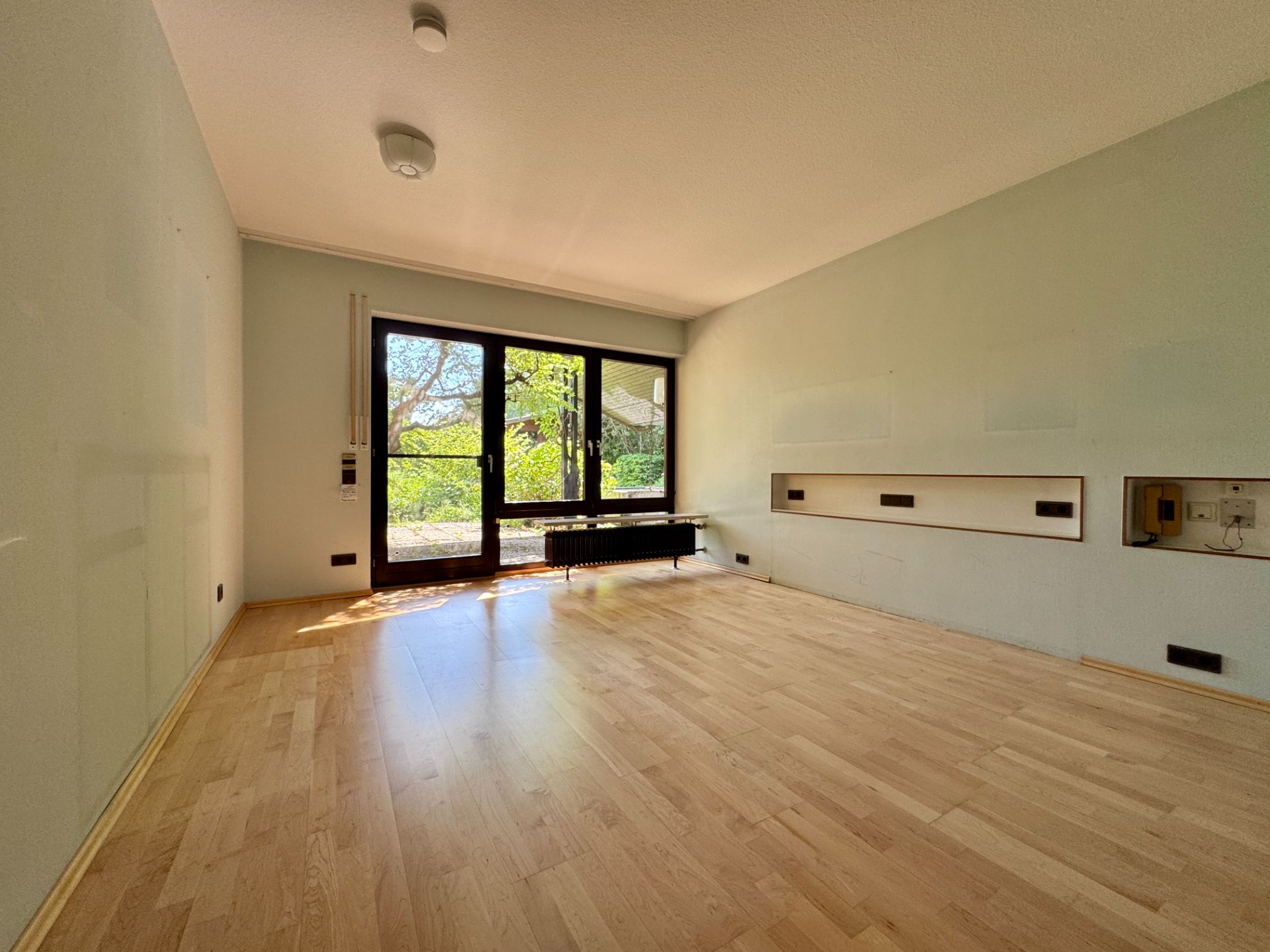 Leeres Zimmer mit hellem Holzboden und großen Fenstern mit Blick ins Grüne.