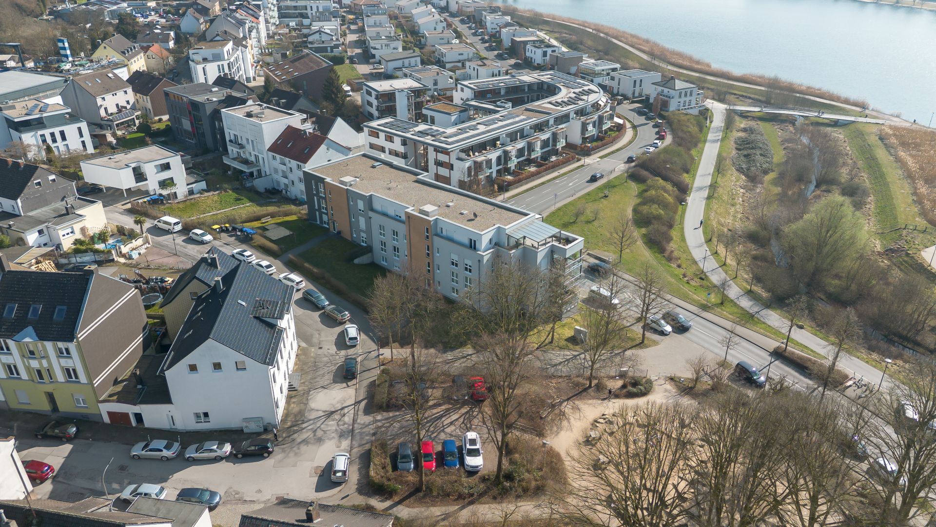 Luftaufnahme von Wohngebäuden, Straßen und geparkten Autos neben einem Flussufer mit Bäumen.
