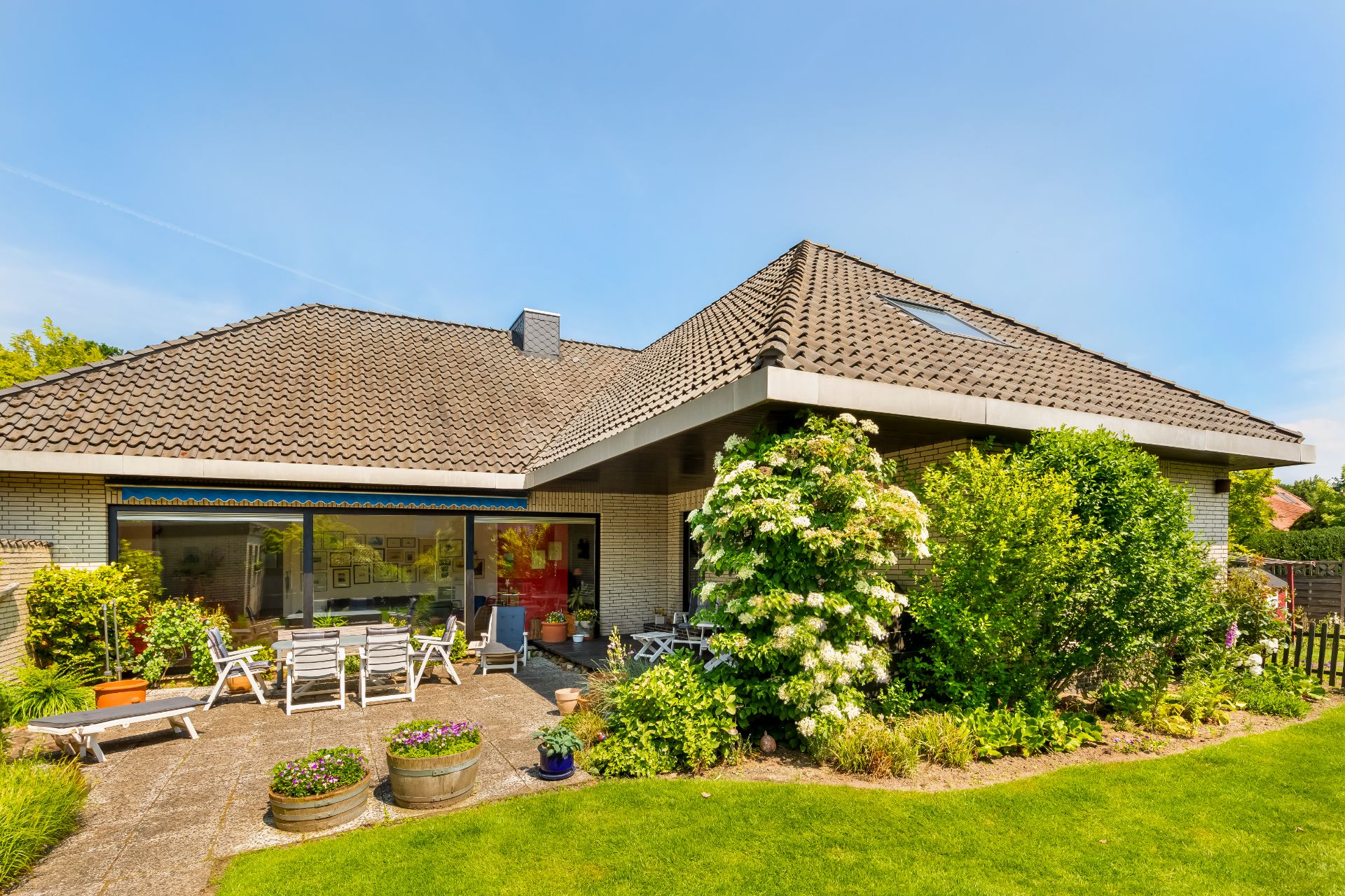 Einfamilienhaus mit Terrasse, Gartenmöbeln, Pflanzen und grünem Rasen bei sonnigem Wetter.