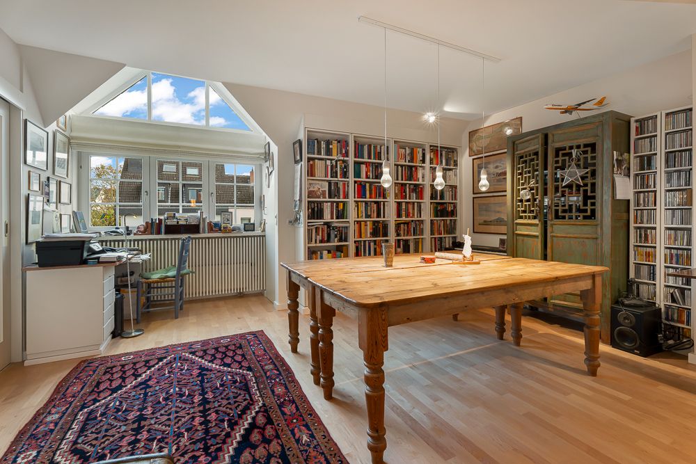 Heller Raum mit großem Holztisch, Bücherregalen, Teppich und Schreibtisch an einem Fenster.