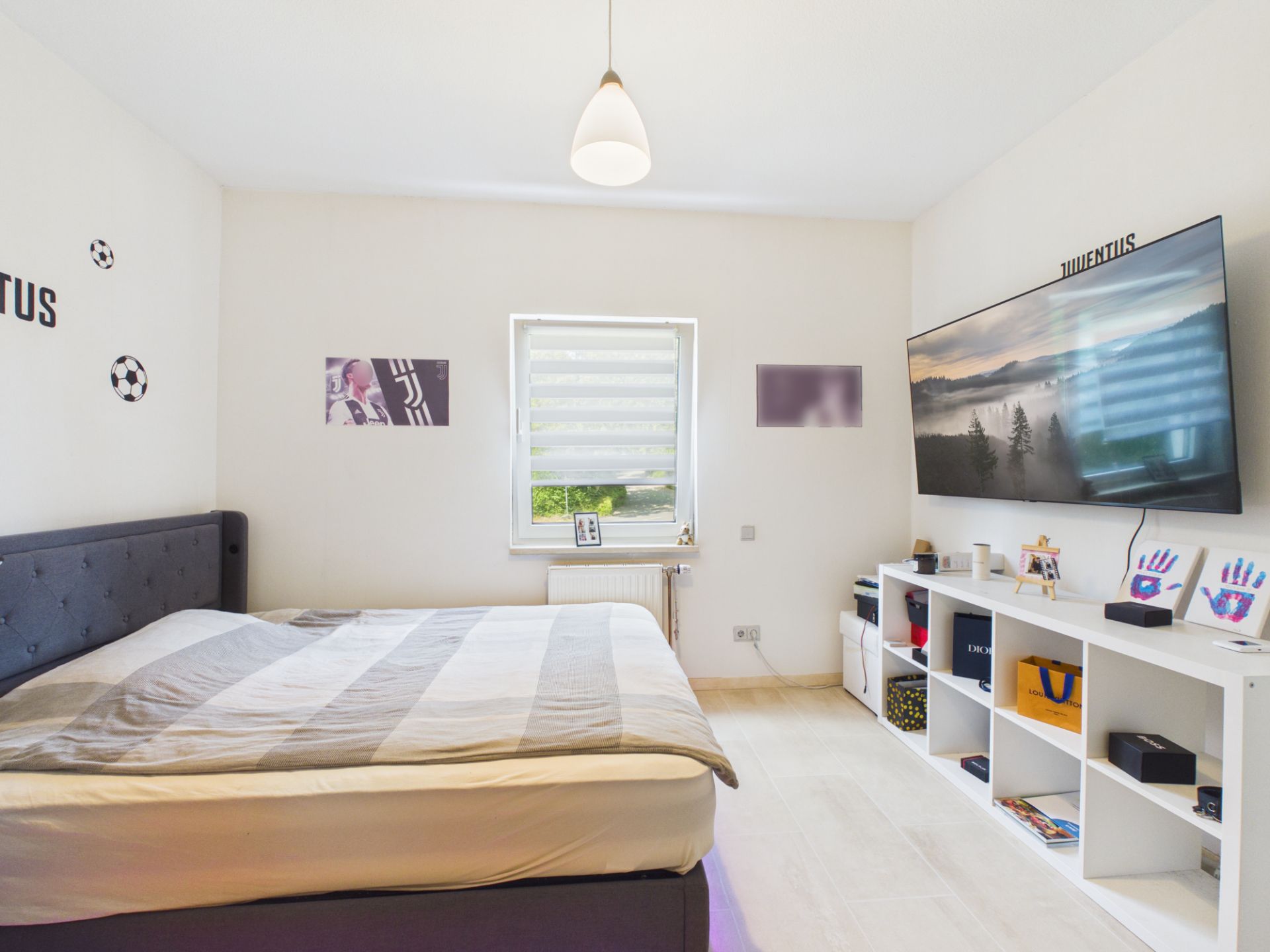 Modernes Schlafzimmer mit großem Bett, Wandfernseher und Regal vor einem Fenster.