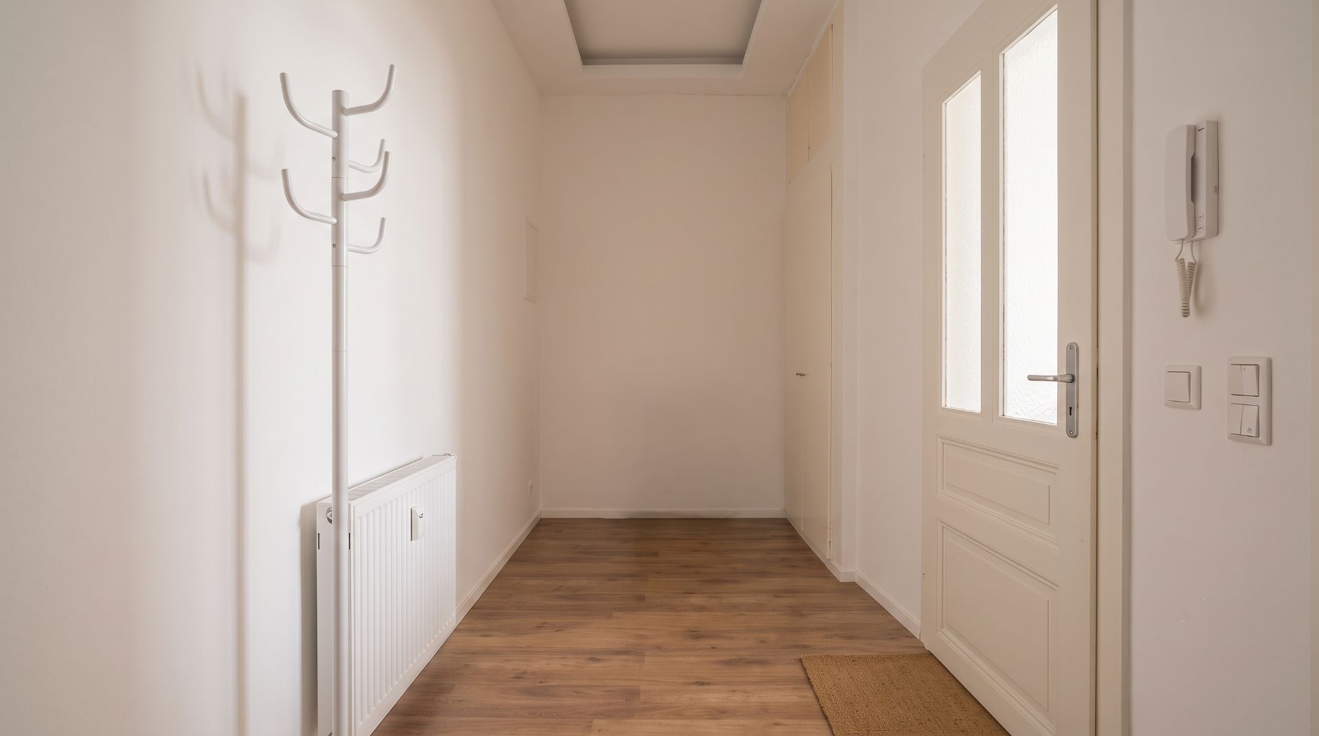 Heller Flur mit Holzboden, Garderobenständer, Heizung und weißer Tür mit Glasfenstern.