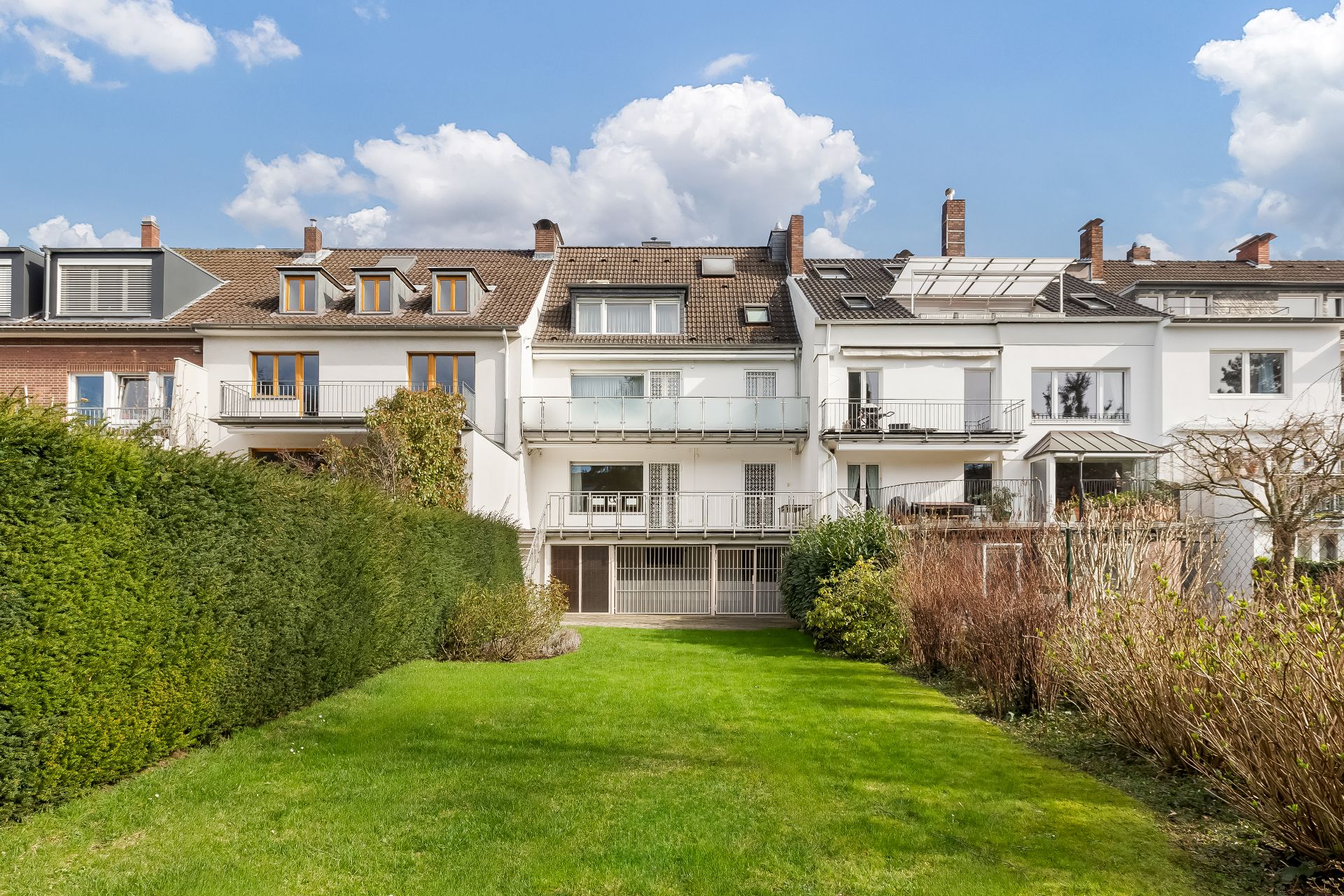 Mehrfamilienhaus mit Balkonen und großem Garten im Vordergrund, umgeben von Hecken und Sträuchern.