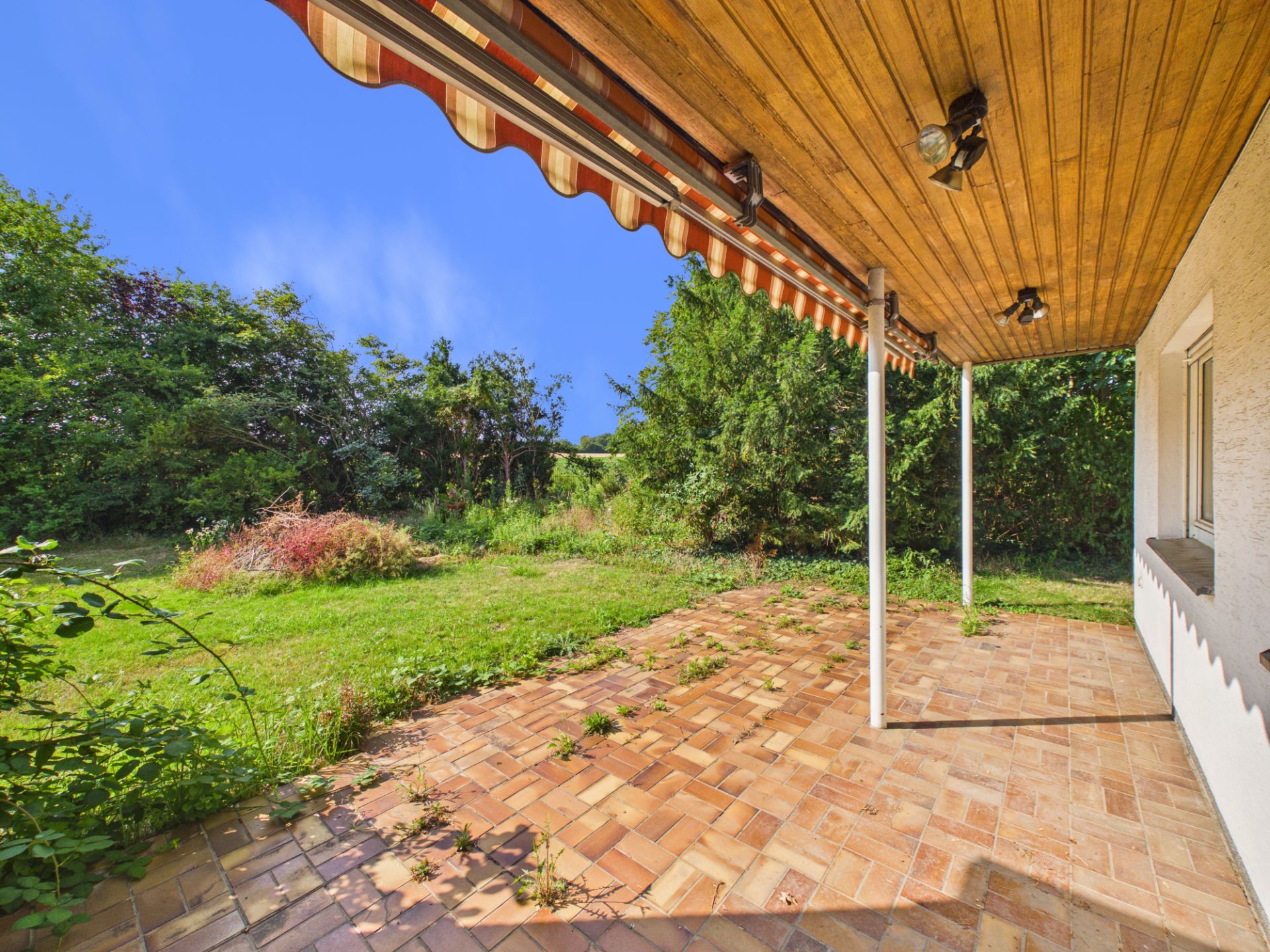Überdachte Terrasse mit Fliesenboden und Blick auf einen großen Garten mit Bäumen.