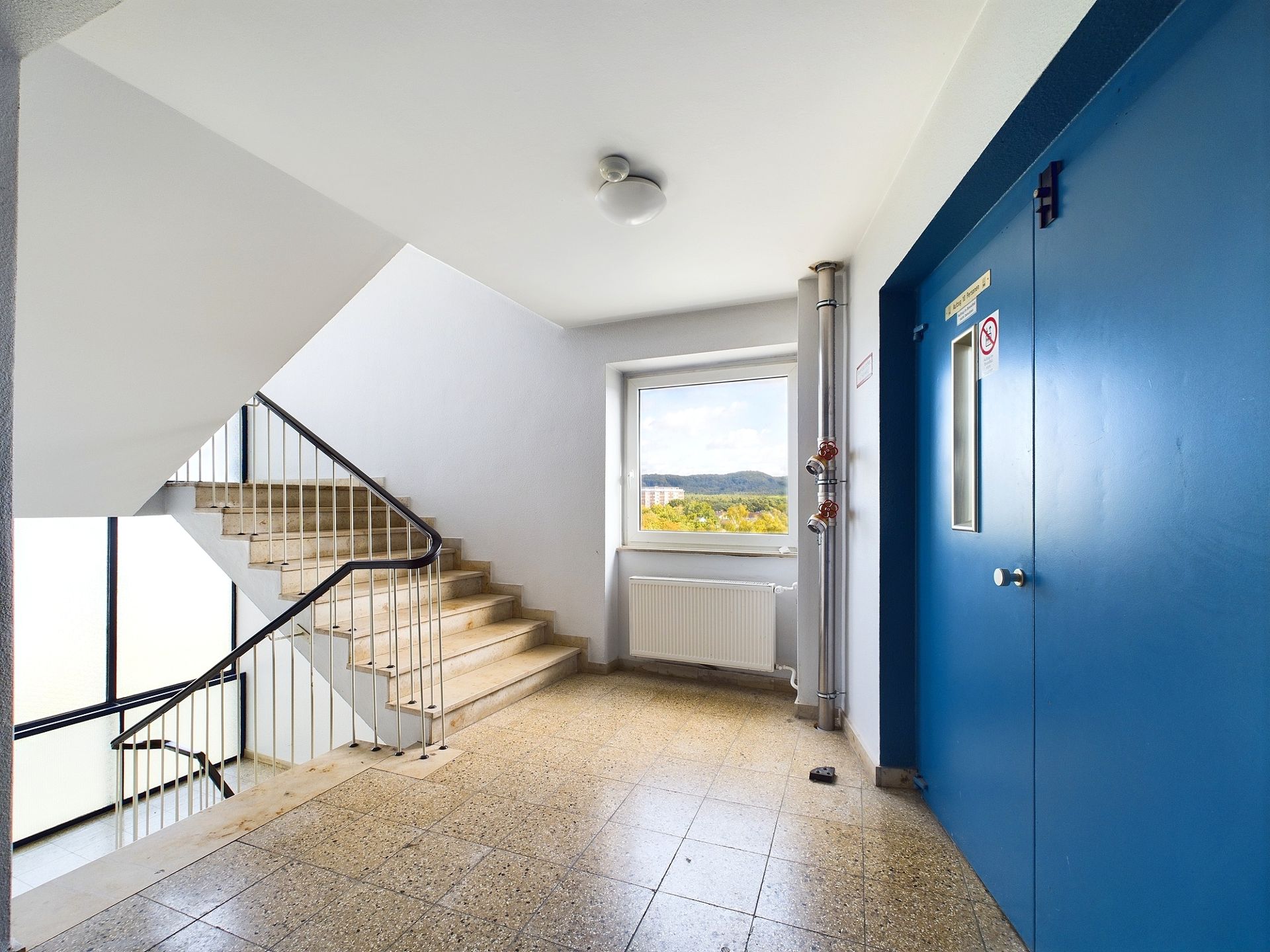 Treppenhaus mit blauen Aufzugtüren, Fenster und Geländer, Fliesenboden.