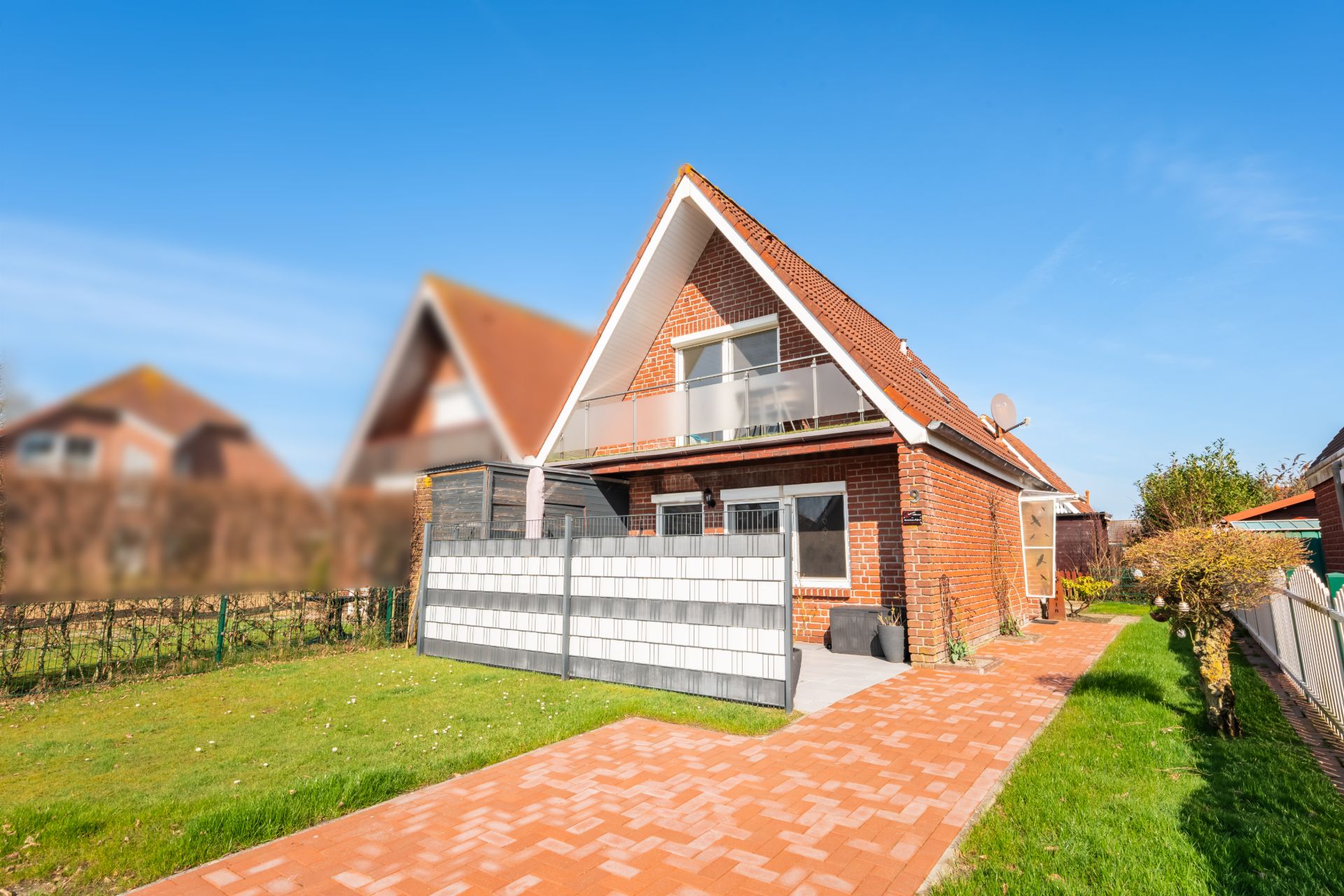 Rotes Backsteinhaus mit Spitzdach, Balkon und kleinem Garten unter blauem Himmel.