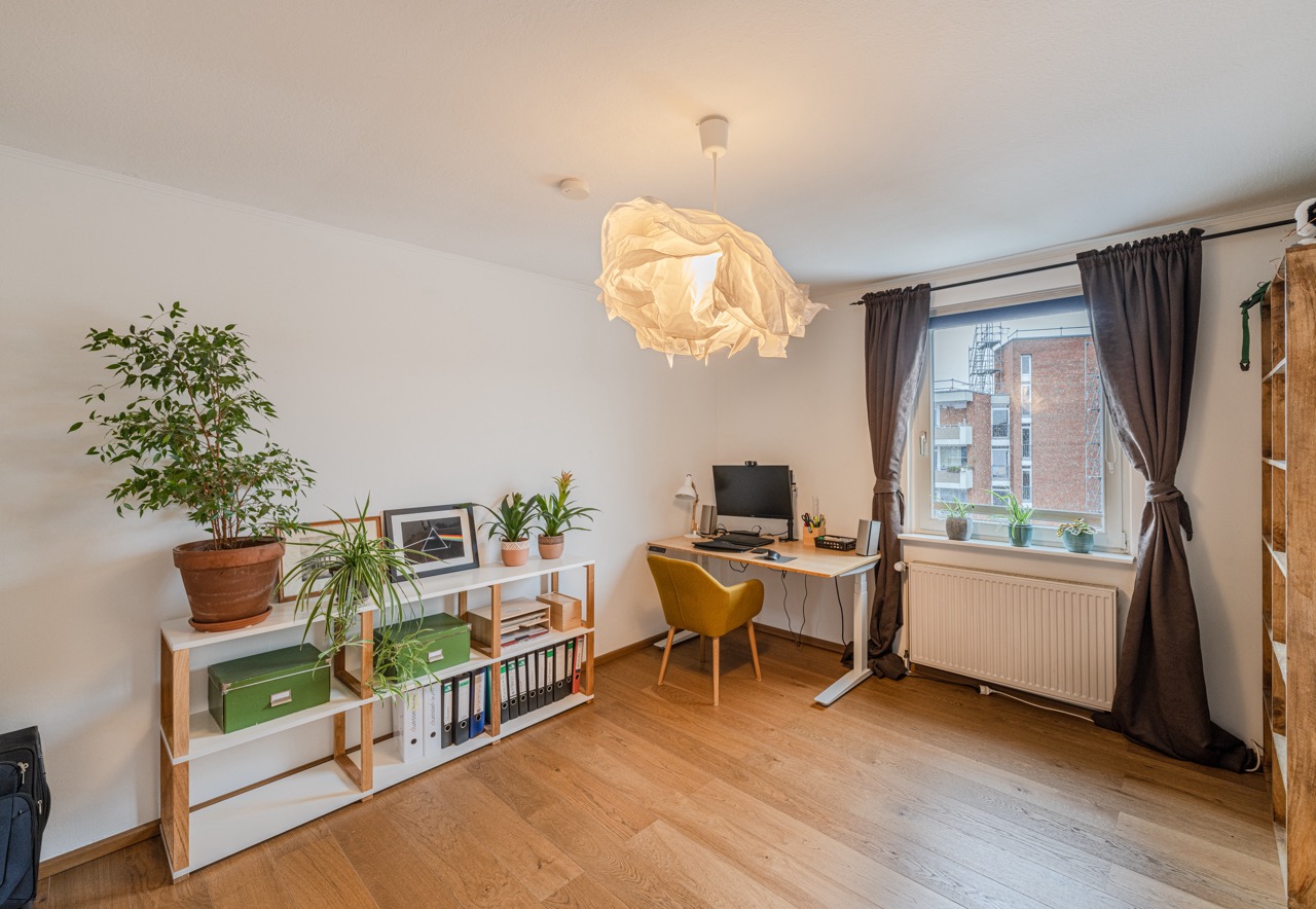 Helles Arbeitszimmer mit Schreibtisch, Regal, Pflanzen und großem Fenster mit Vorhängen.