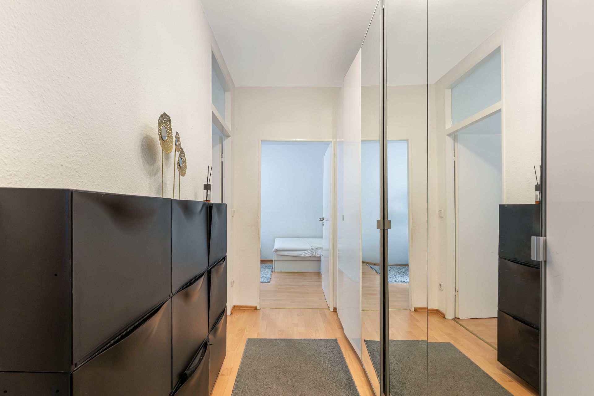 Heller Flur mit schwarzen Kommoden, Spiegelwand und Holzboden, Blick in ein weiteres Zimmer.