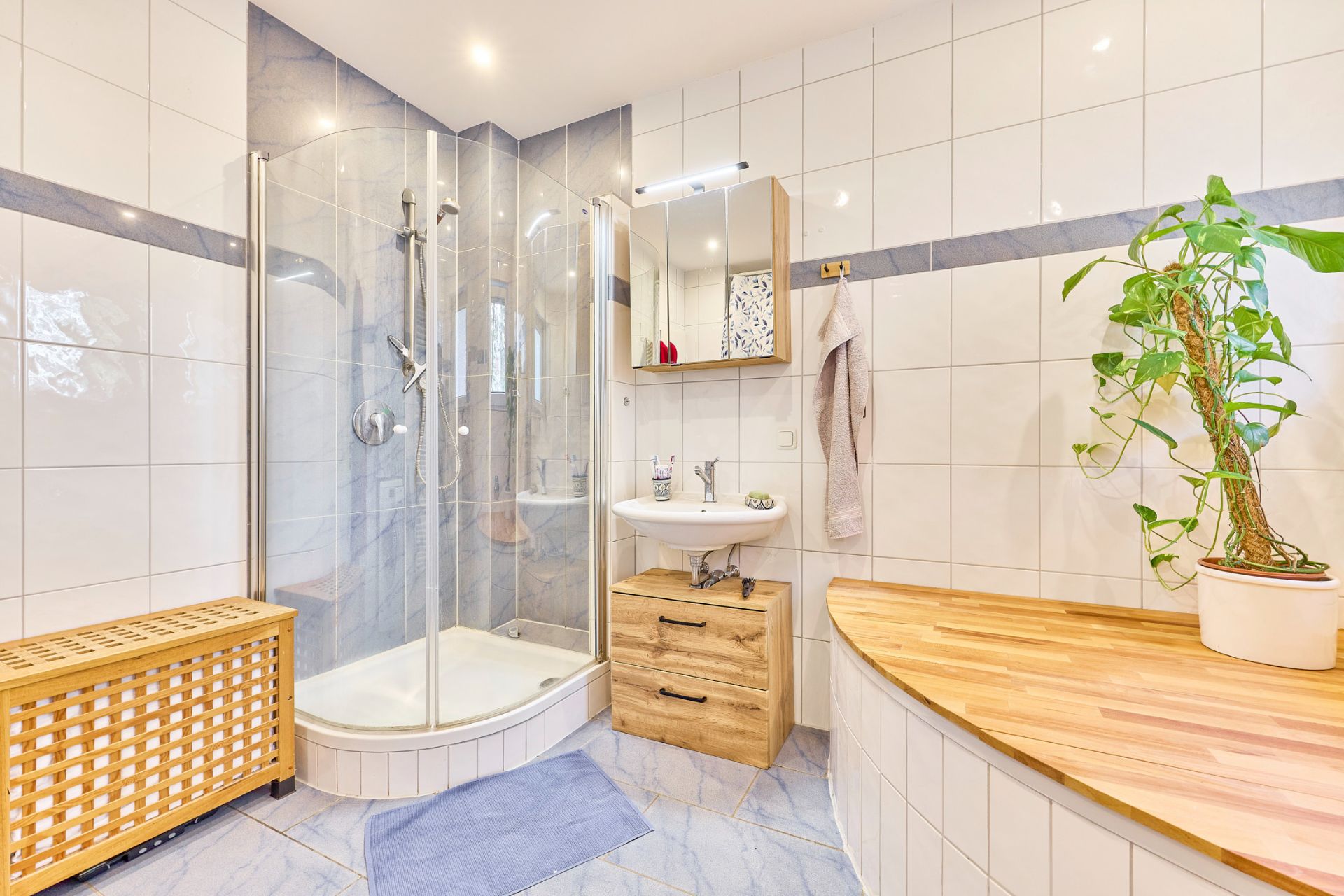 Modernes Badezimmer mit Eckdusche, Waschbecken, großem Spiegel, Holzablage und Zimmerpflanze.