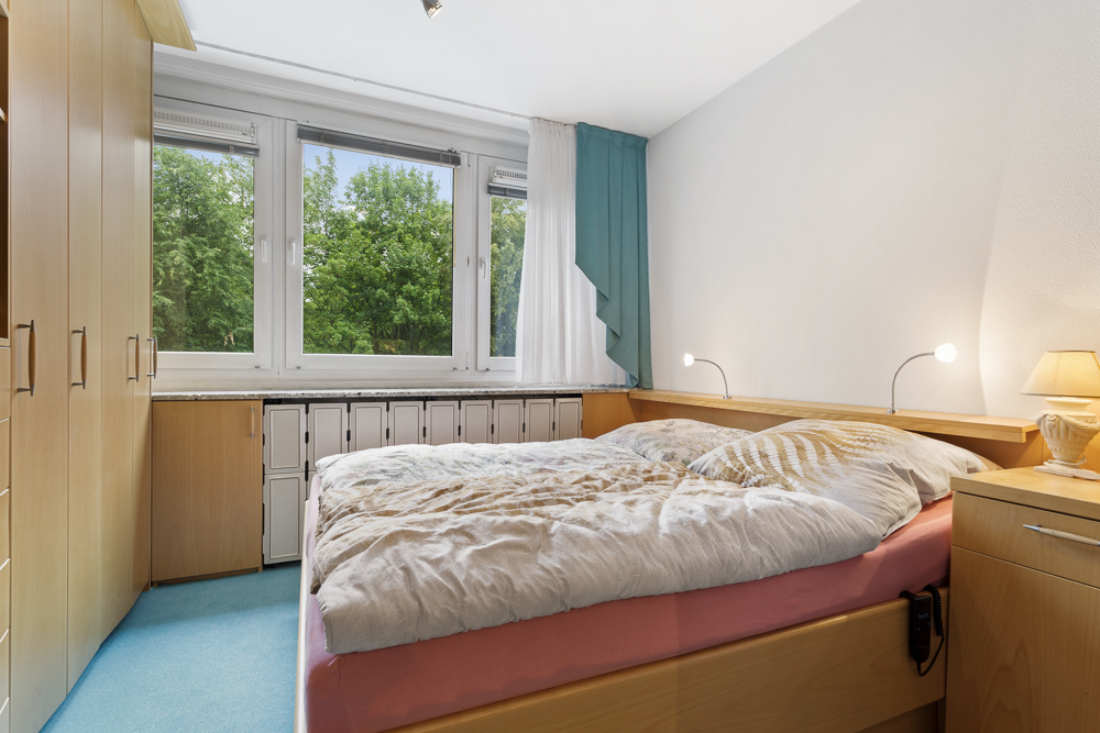 Helles Schlafzimmer mit großem Fenster, Doppelbett, Schränken und blauer Teppichboden.