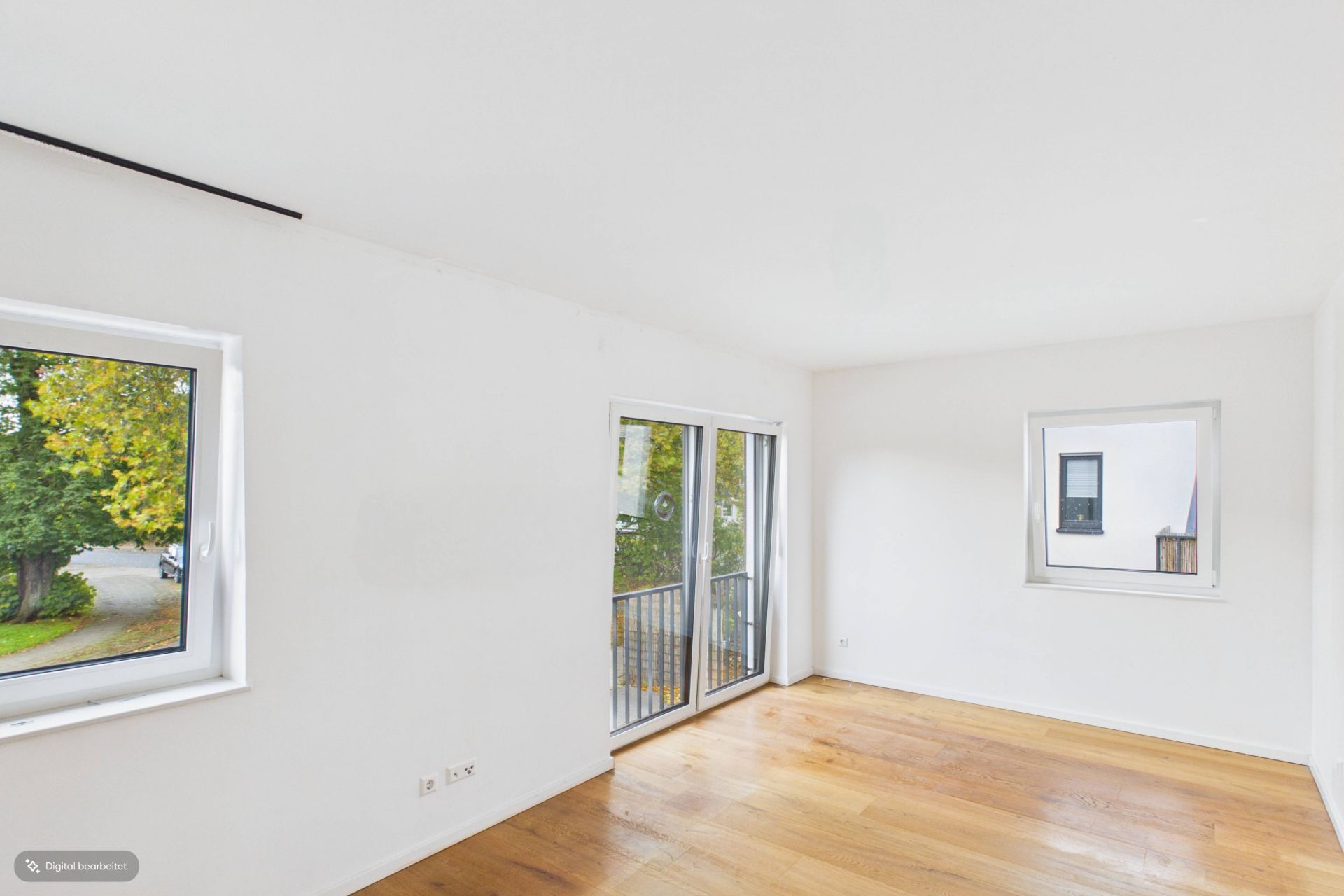 Helles, leeres Zimmer mit Holzfußboden, zwei Fenstern und einer Glastür zum Balkon.