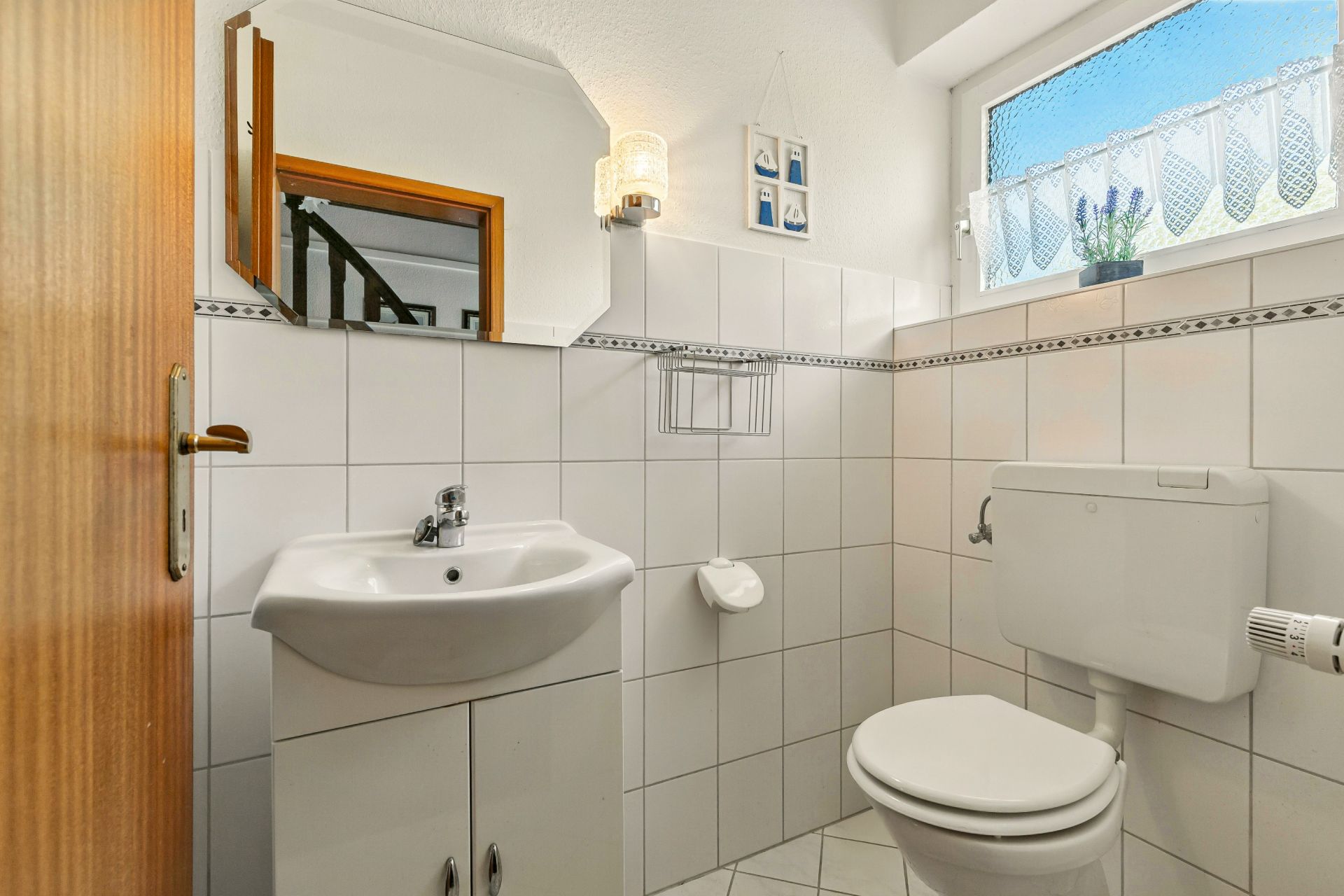 Kleines, helles Bad mit Waschbecken, Spiegel, Toilette und Fenster mit Gardine.