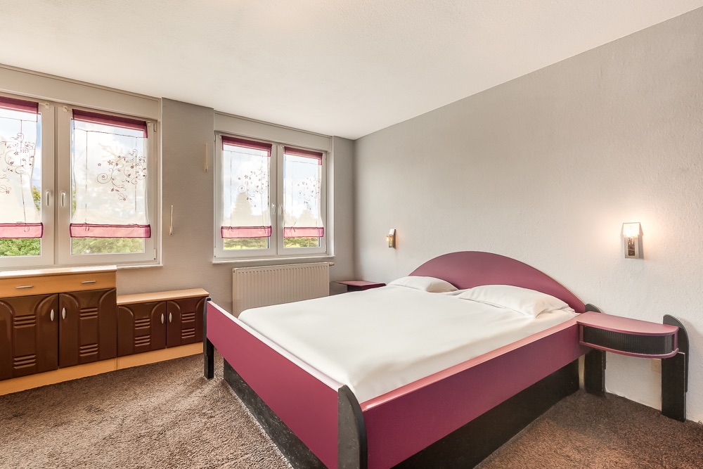Schlafzimmer mit großem Bett, drei Fenstern mit Rollos und braunen Kommoden auf Teppichboden.