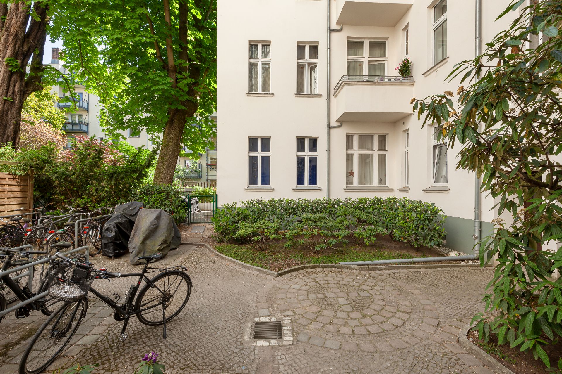 Gepflasterter Innenhof mit Fahrrädern, Sträuchern und einem weißen Wohnhaus im Hintergrund.