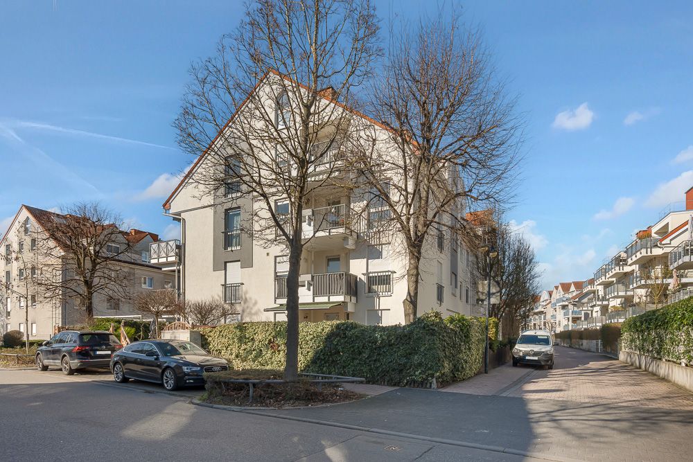 Mehrfamilienhaus mit Balkonen, Bäumen und geparkten Autos an einer ruhigen Straße bei sonnigem Wetter.