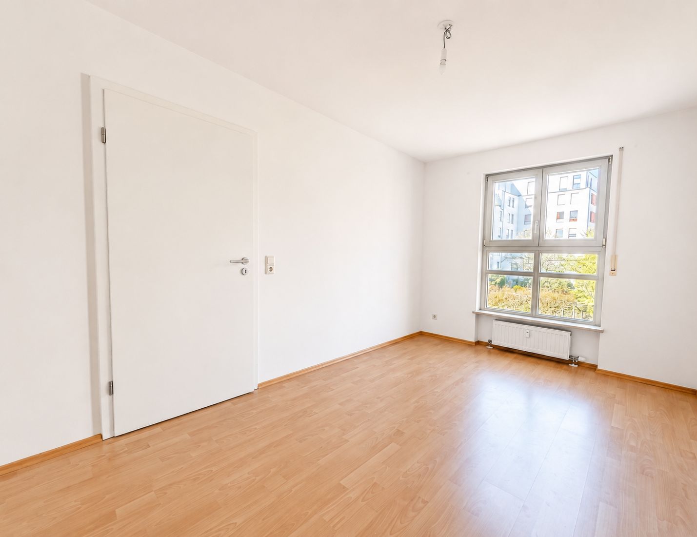 Helles, leeres Zimmer mit Holzfußboden, Fenster und weißer Tür.