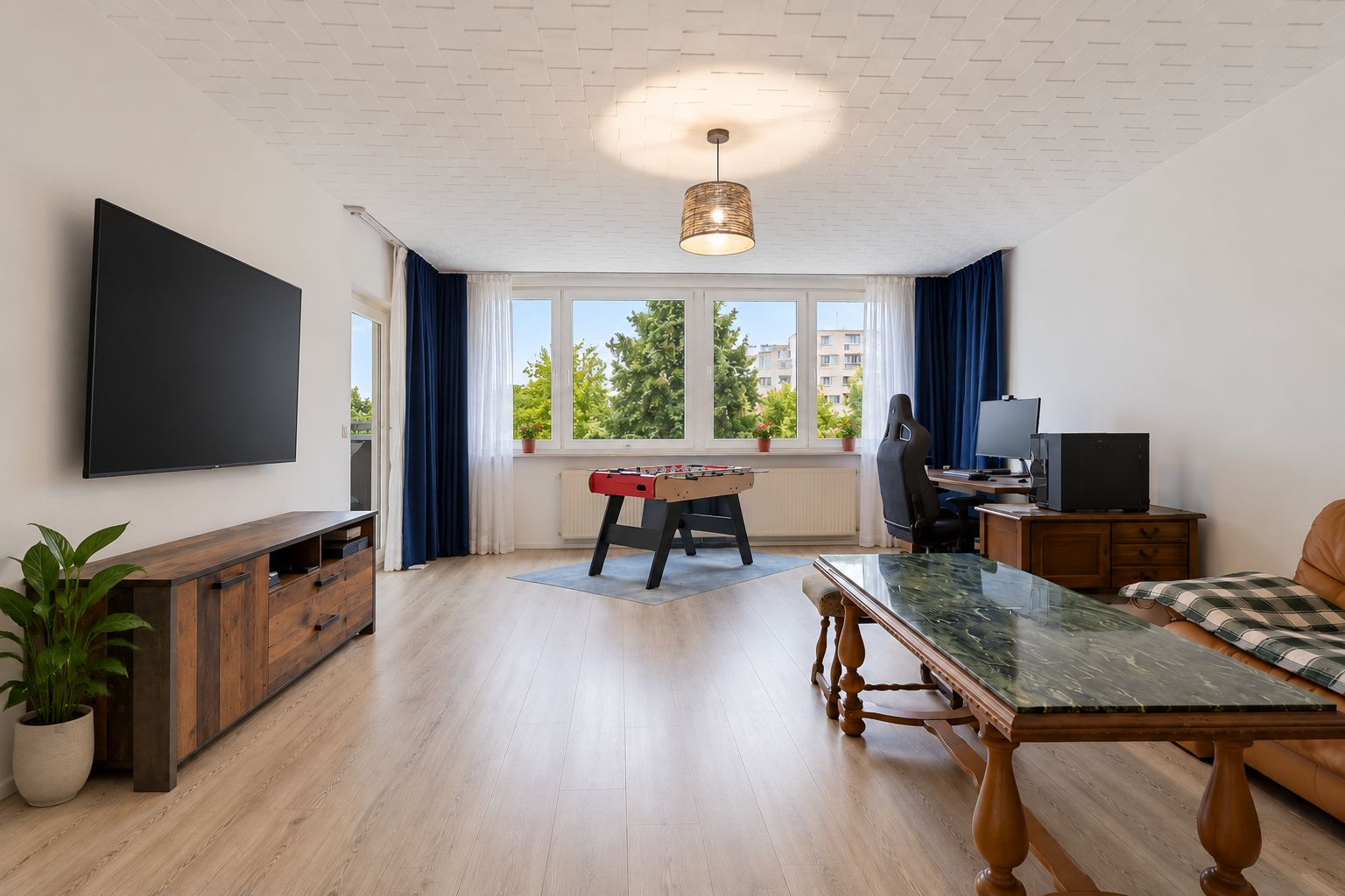 Helles Wohnzimmer mit großem Fenster, Kickertisch, Schreibtisch, Sofa und Fernseher an der Wand.