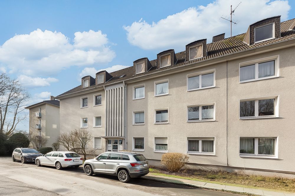 Mehrfamilienhaus mit drei Etagen, Dachfenstern und parkenden Autos davor.