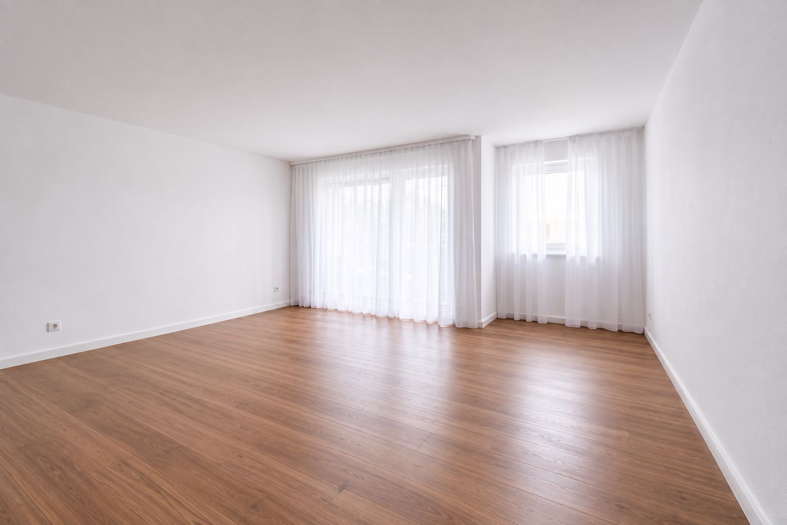 Leerer, heller Raum mit Holzfußboden und großen Fenstern mit weißen Gardinen.