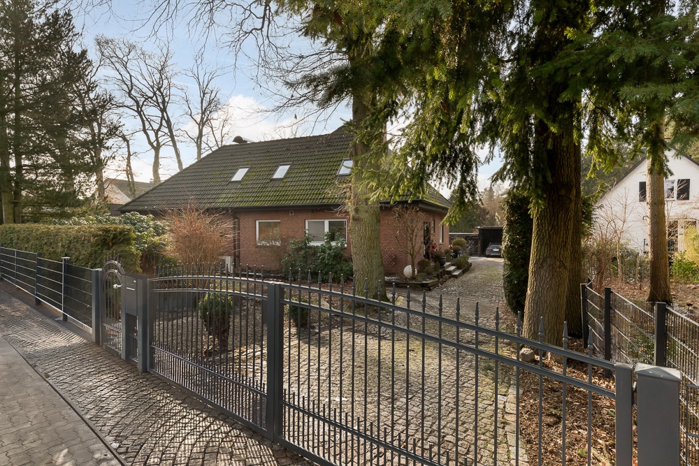 Backsteinwohnhaus mit grünem Dach, Garten und Metallzaun, große Bäume im Vordergrund.