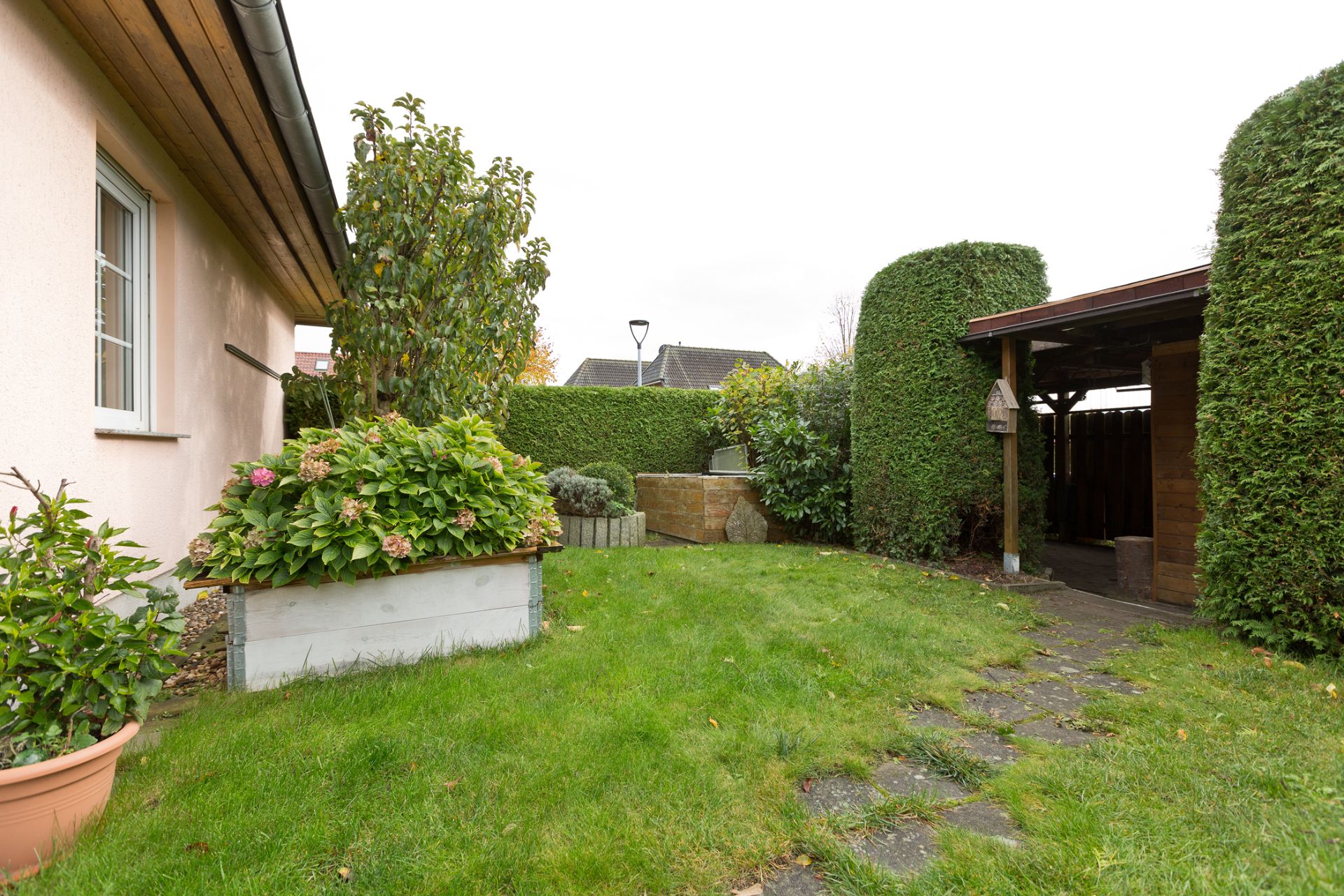 Grüner Garten mit Rasen, Sträuchern, Hecken und einem überdachten Holzbereich an einem Wohnhaus.