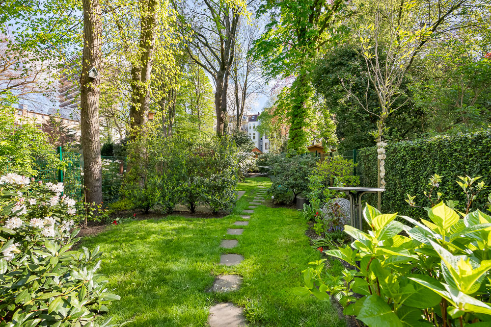 Gepflegter Garten mit grünem Rasen, Blumen, Bäumen und Trittsteinen in der Mitte.