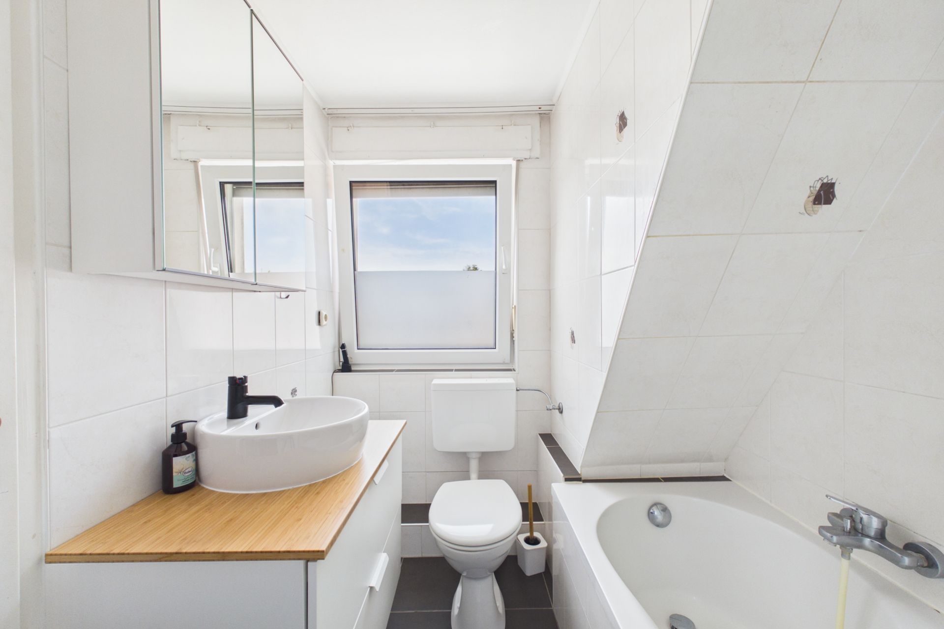 Helles Badezimmer mit Waschbecken, WC, Badewanne und zwei Fenstern.