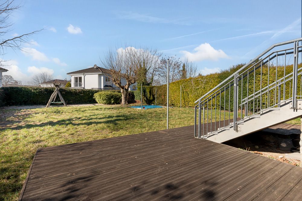 Garten mit Holzterrasse, Metalltreppe, Schaukel und Rasen, umgeben von Büschen.