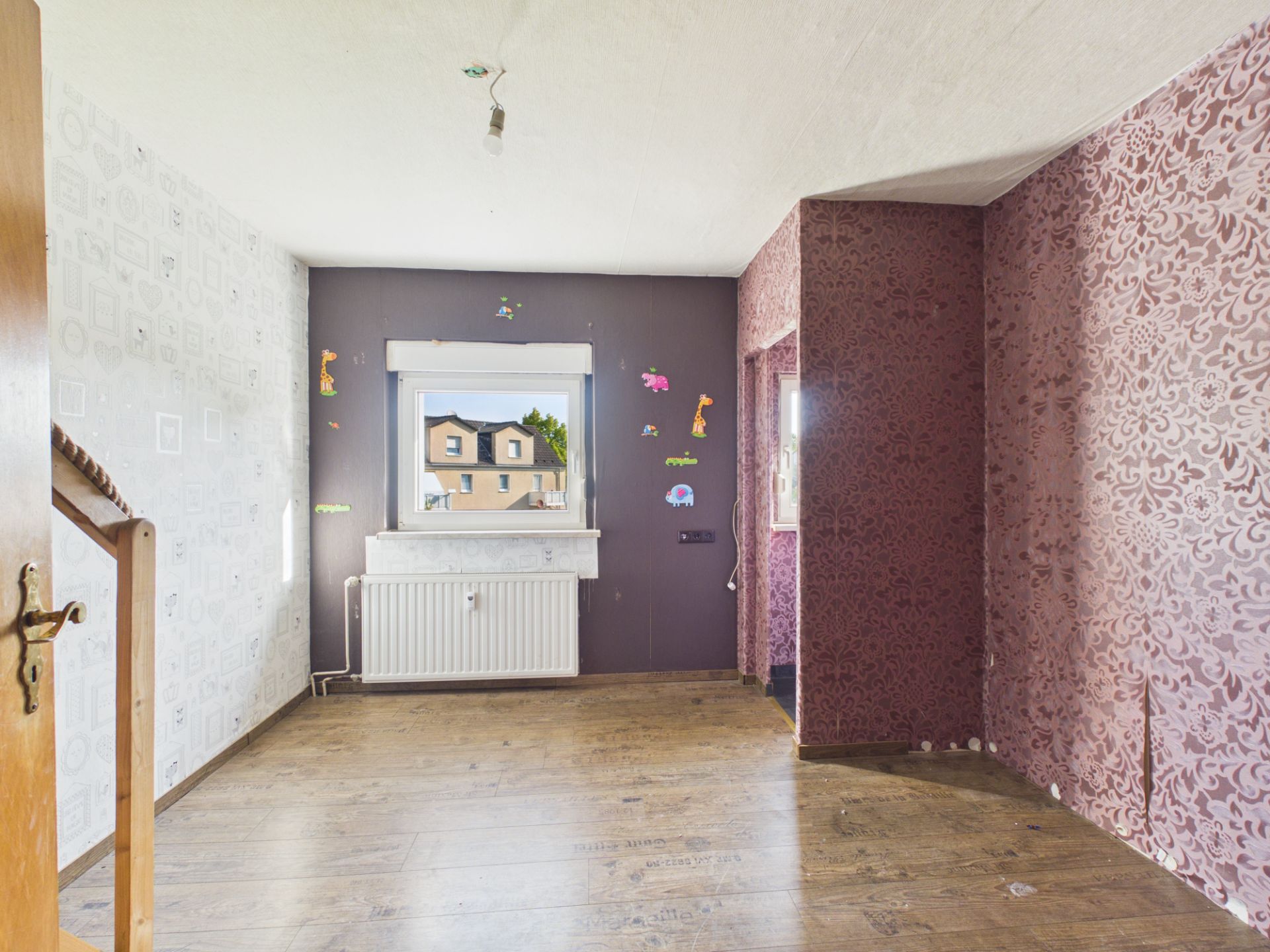 Leeres Zimmer mit Holzfußboden, gemusterten Tapeten und Wandaufklebern, Fenster mit Blick nach draußen.