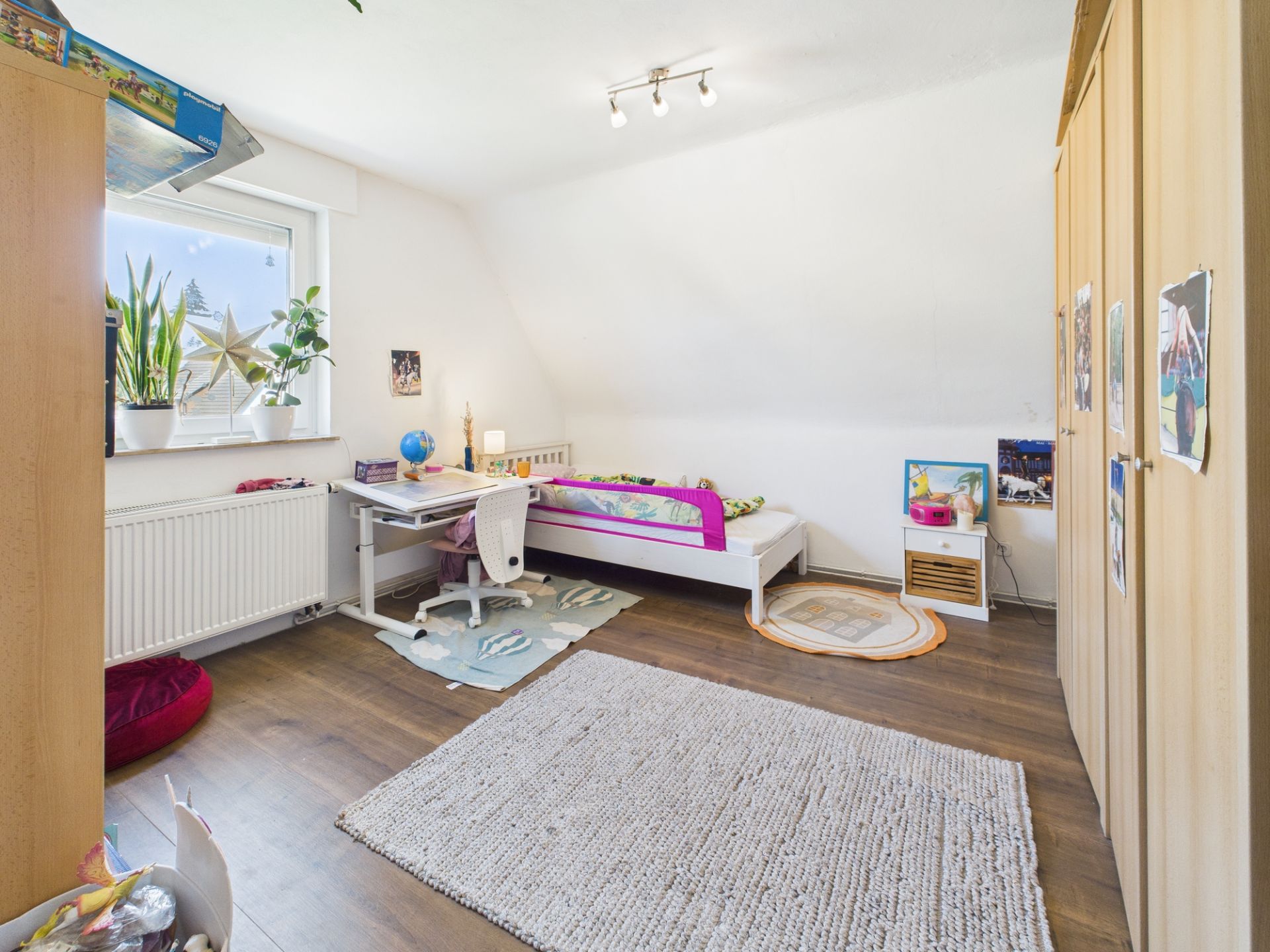 Helles Kinderzimmer mit Bett, Schreibtisch, Teppich und Schrank, Holzfußboden und Fenstern mit Pflanzen.